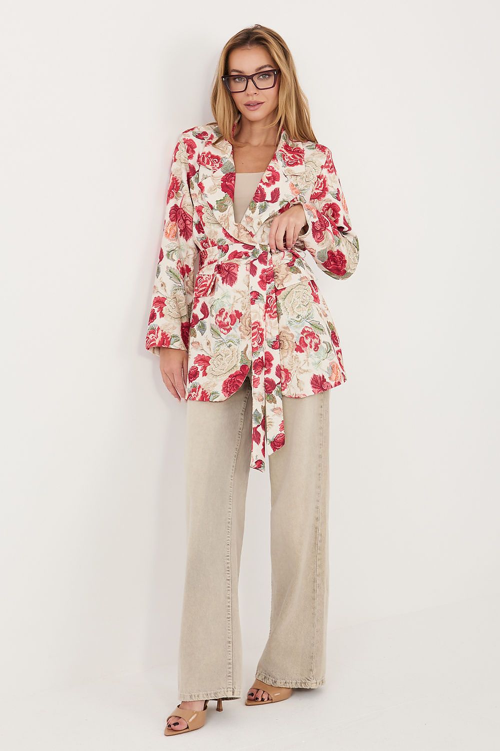 A woman modeling a long-sleeve floral wrap blazer with a red and pink rose pattern on a cream background, paired with beige wide-leg jeans and brown heeled sandals, against a plain white studio background.