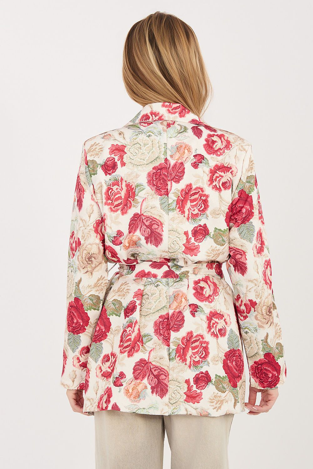 A woman shown from the back wearing a long-sleeve belted jacket with a red and pink rose floral print on a cream background, paired with beige trousers, against a light gray studio background.