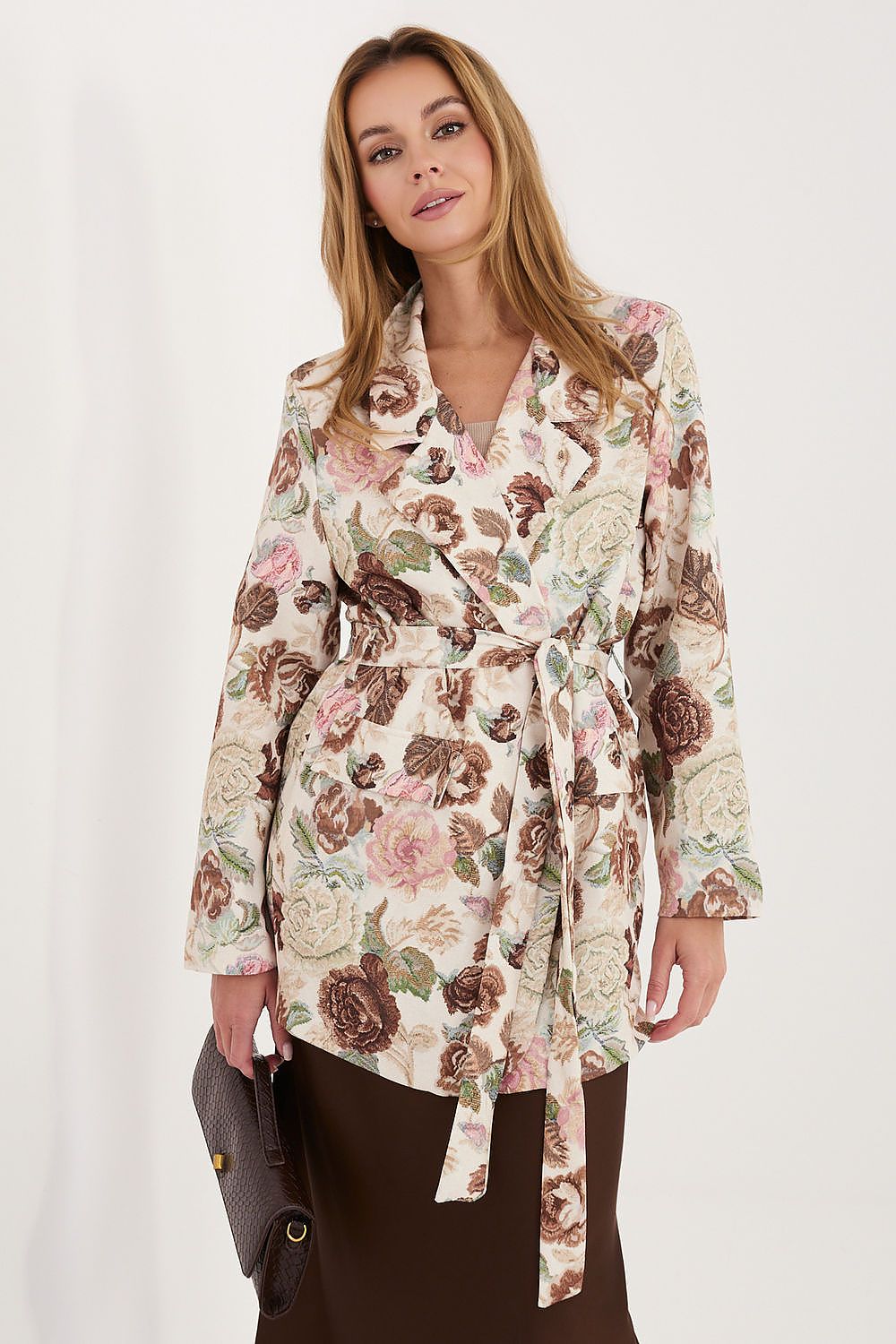 A woman wearing a long-sleeve floral wrap jacket with a tie waist, featuring a cream base with pink, brown, and green botanical print, paired with dark brown bottoms and holding a dark brown clutch.