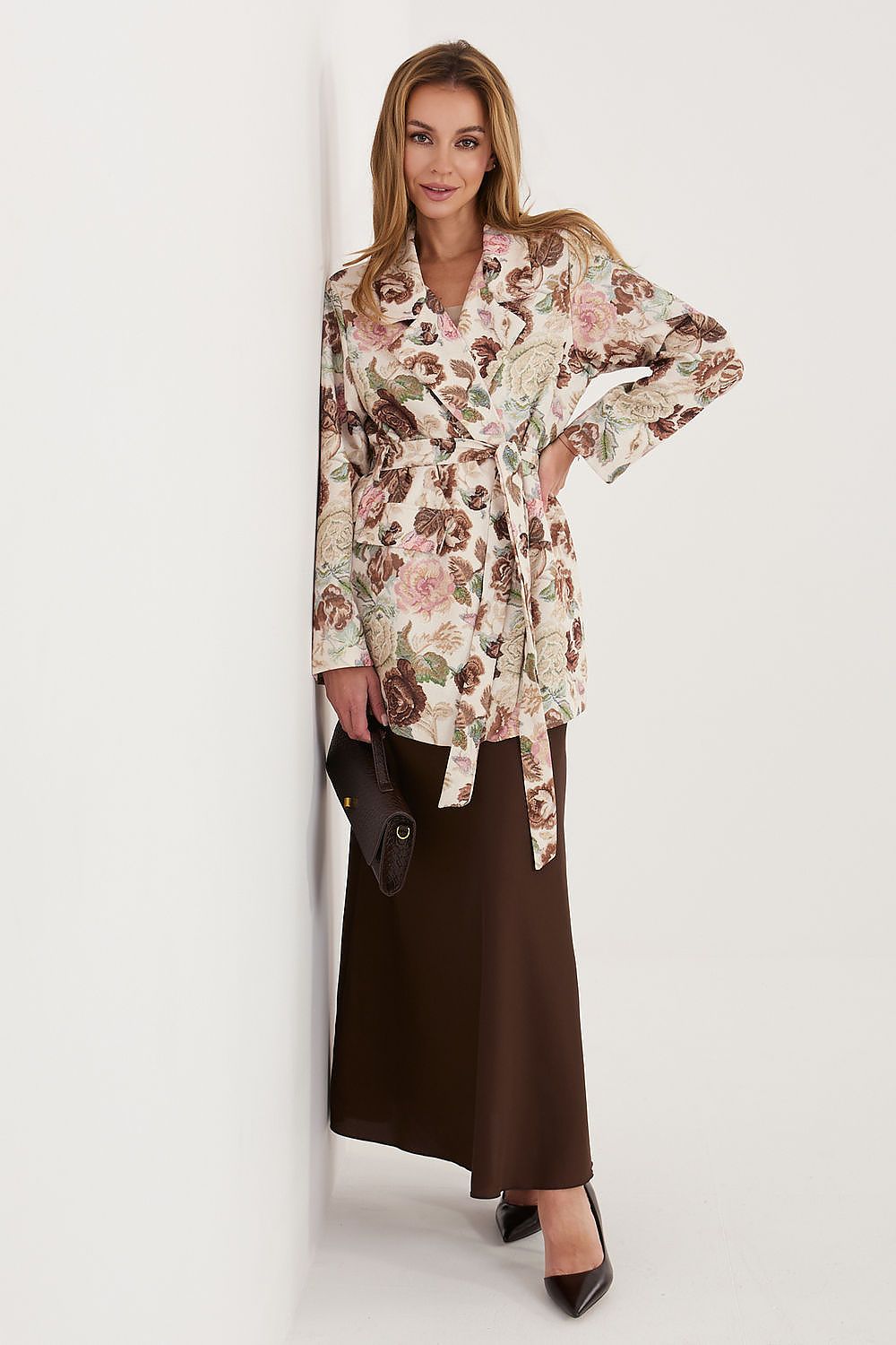 A woman models a long-sleeve floral wrap jacket with a tie waist, paired with a dark brown maxi skirt and black heels, standing against a plain white wall.