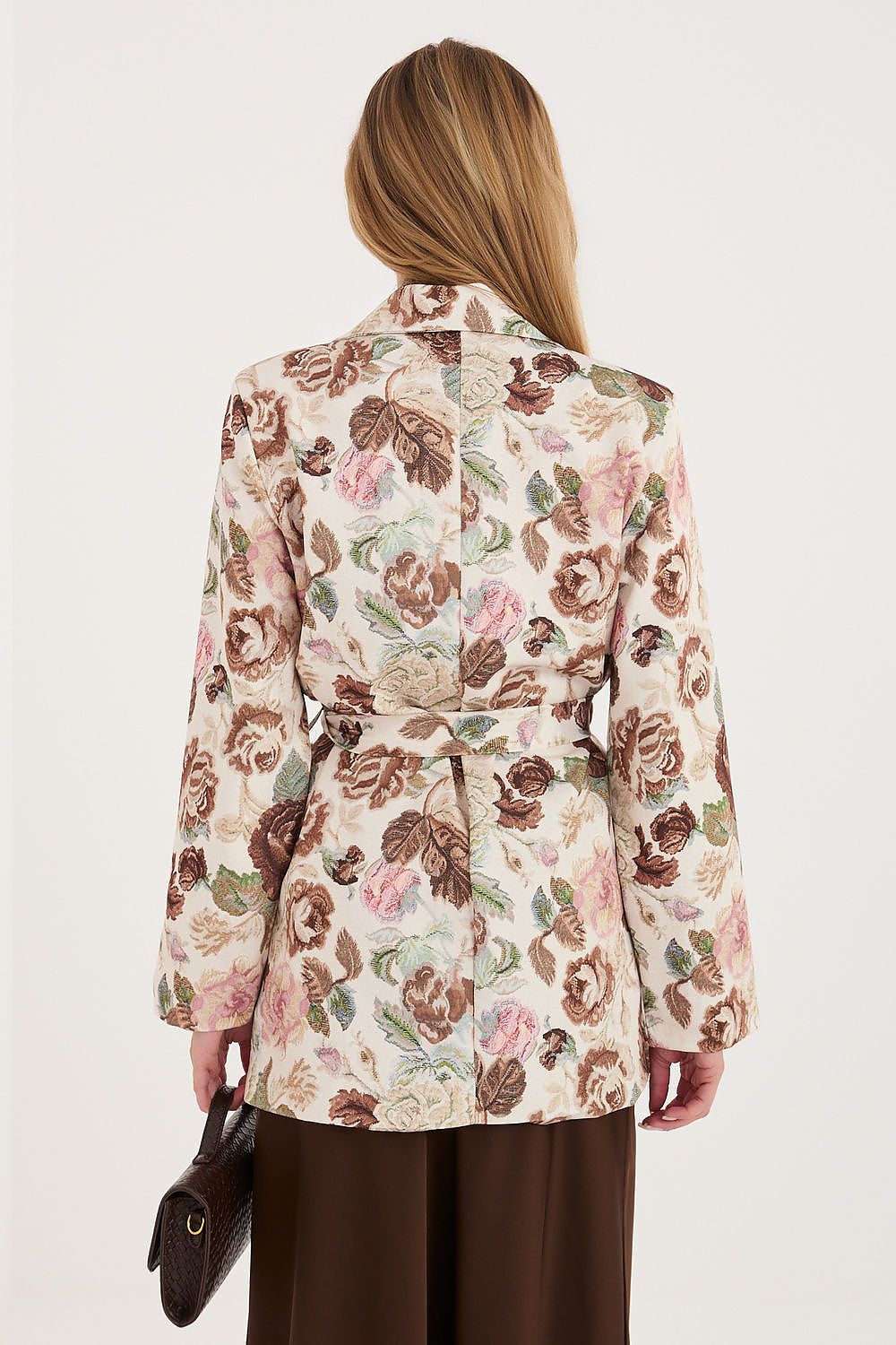 A woman shown from the back wearing a long-sleeve belted floral jacket with a cream base and pink, brown, and green botanical print, paired with dark brown pants against a plain light background.