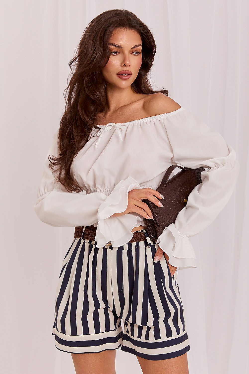 A woman modeling a white off-the-shoulder blouse with long ruffled sleeves and navy-and-white vertically striped shorts, holding a dark brown clutch against a light draped fabric backdrop.