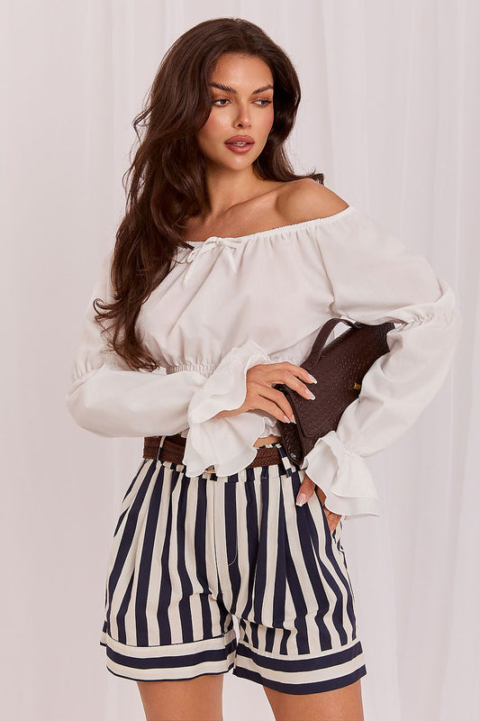 A woman modeling a white off-the-shoulder blouse with long ruffled sleeves and navy-and-white vertically striped shorts, holding a dark brown clutch against a light draped fabric backdrop.
