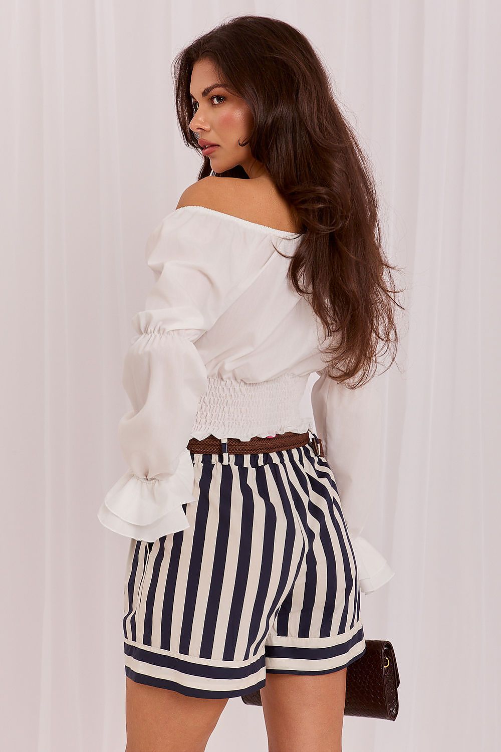 A woman wearing a white off-the-shoulder blouse with long ruffled sleeves and navy-and-white vertically striped shorts, photographed against a light draped fabric backdrop.