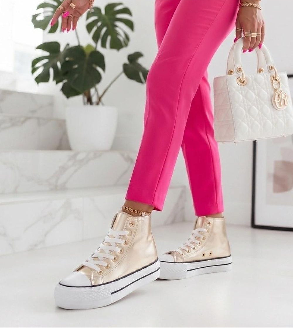 A person wearing bright pink pants and metallic gold high-top platform sneakers with white soles, holding a white quilted handbag in a bright indoor setting with a potted plant and marble steps.
