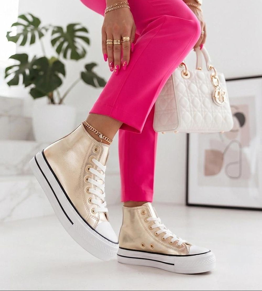 A person wearing metallic gold high-top platform sneakers with white soles, paired with bright pink pants and holding a white quilted handbag in a bright indoor setting.