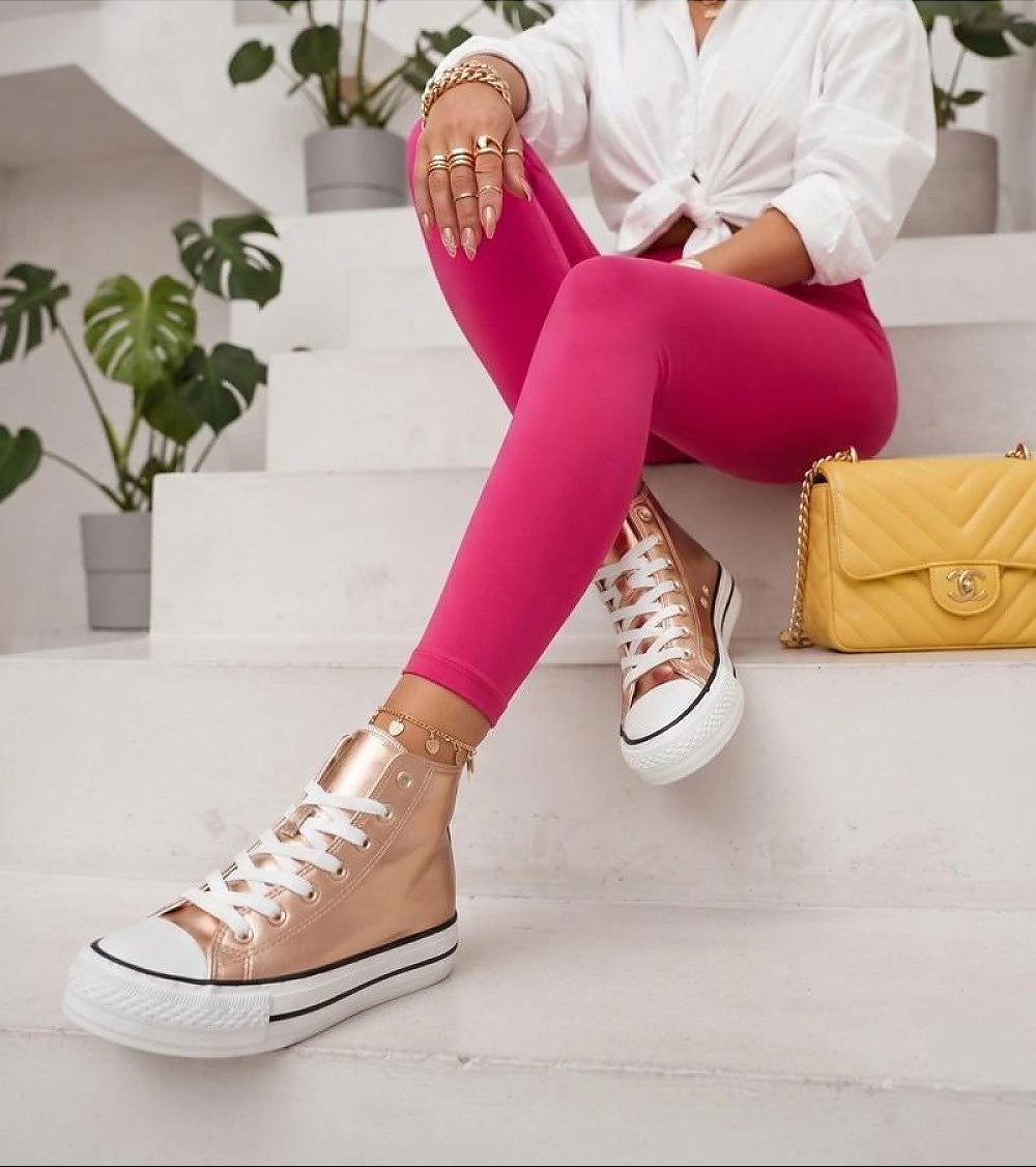 A person seated on white steps wearing bright pink leggings, a white tied blouse, metallic rose-gold high-top sneakers, gold rings, and a yellow quilted handbag.
