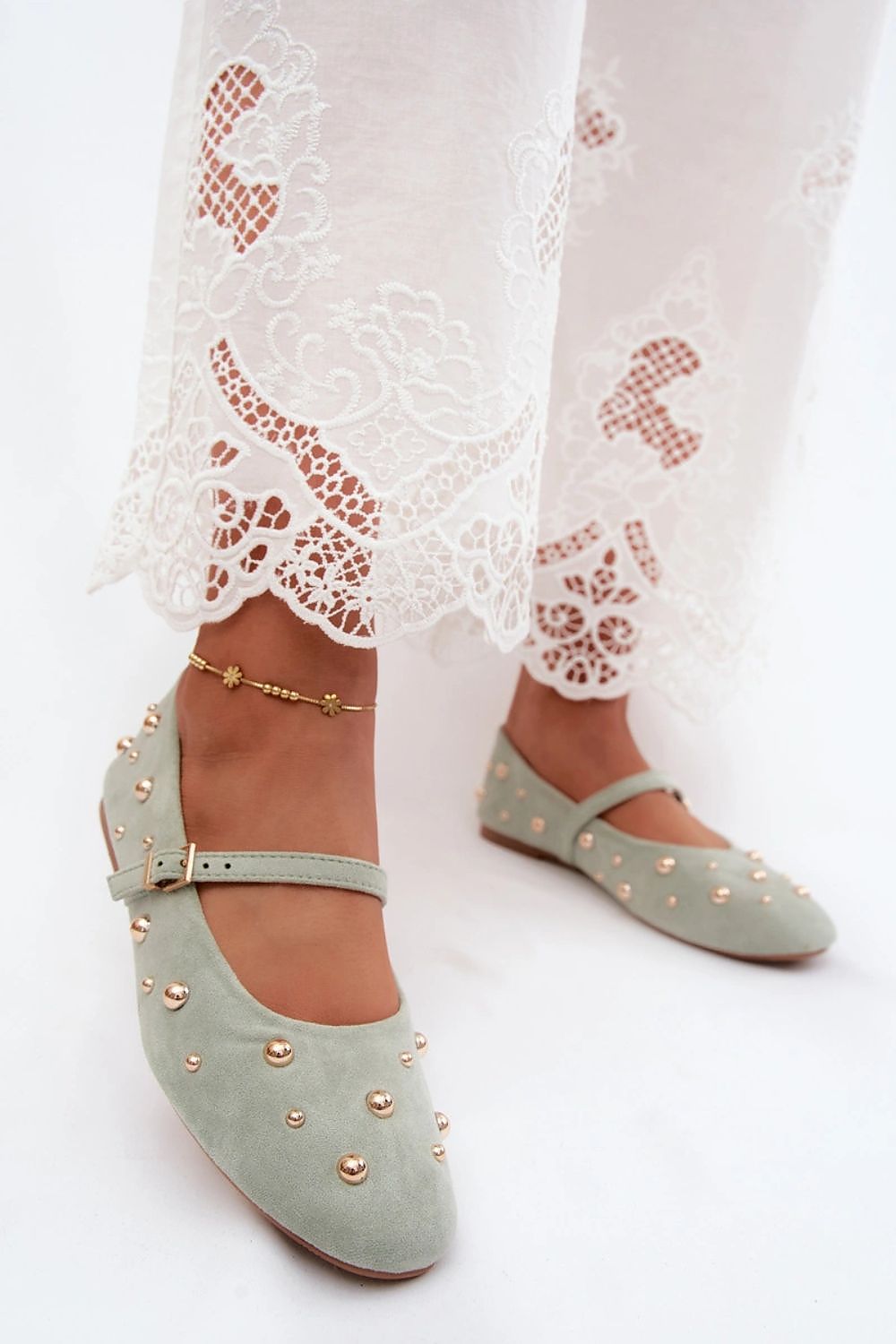 A pair of sage green suede flat shoes with gold-tone studs and ankle straps, shown on a model wearing white lace-trimmed pants against a white background.