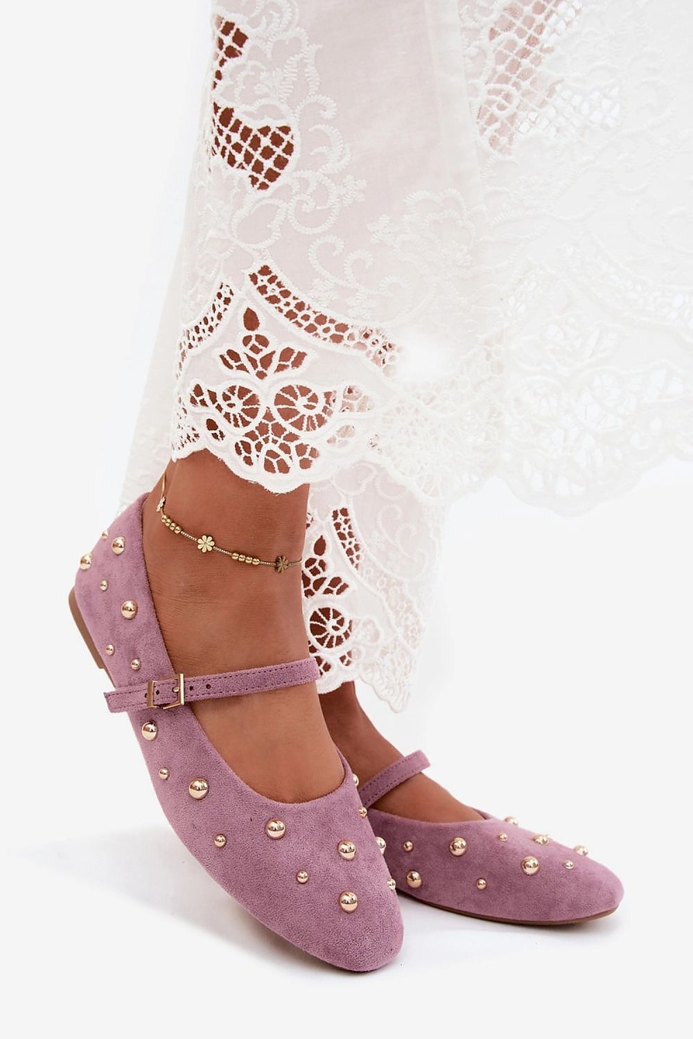 A pair of lavender-purple suede ballet flats decorated with gold-tone studs, shown on a model's feet wearing a white lace dress against a white studio background.