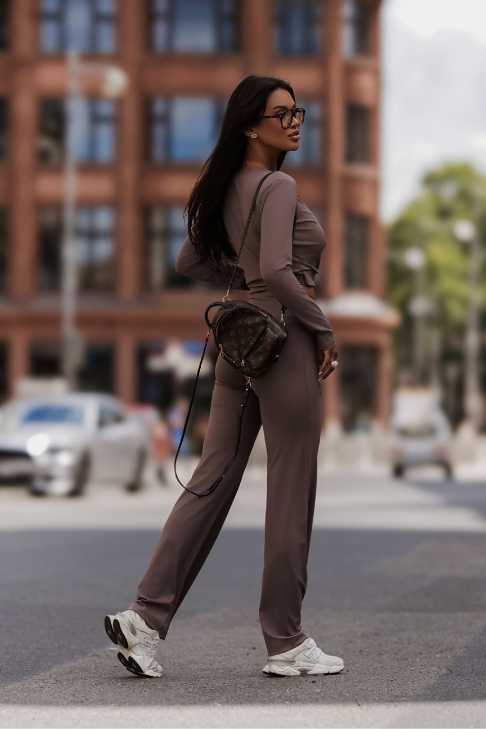 A woman wearing a matching brown long-sleeve top and wide-leg pants set, styled with a dark brown crossbody bag and white sneakers, walking on a city street with a blurred brick building in the background.