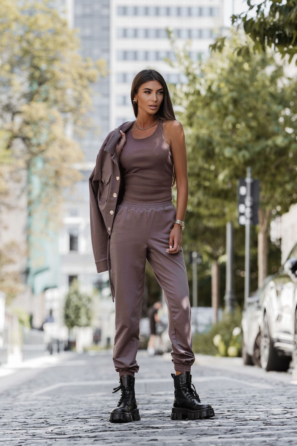 A woman stands on a cobblestone street wearing a matching taupe-brown lounge set consisting of a ribbed tank top, jogger pants, and an open button-up jacket draped over her shoulder, paired with black combat boots.