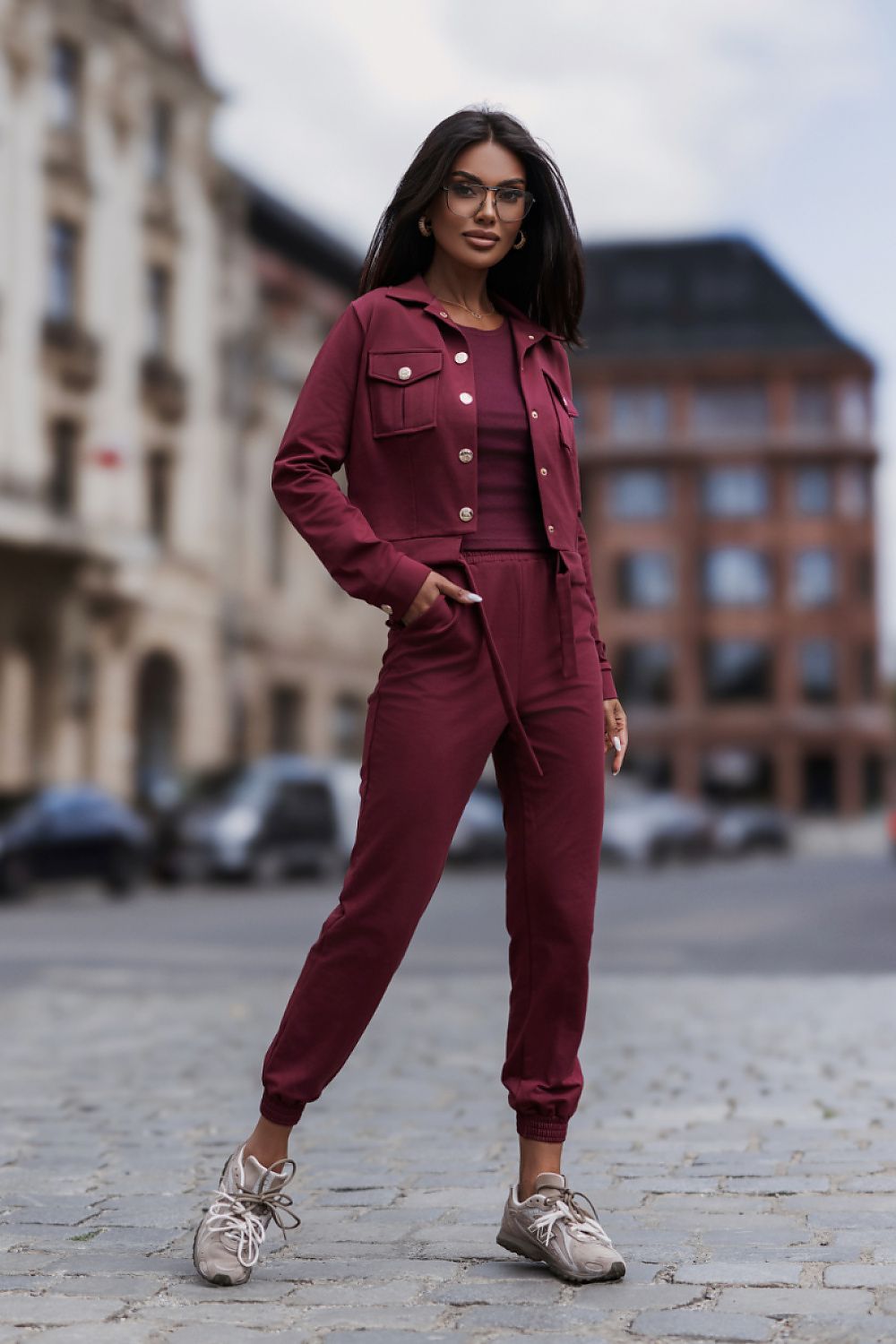 A woman wearing a matching burgundy two-piece tracksuit with a button-front jacket and jogger pants, standing on a cobblestone street with blurred city buildings in the background.