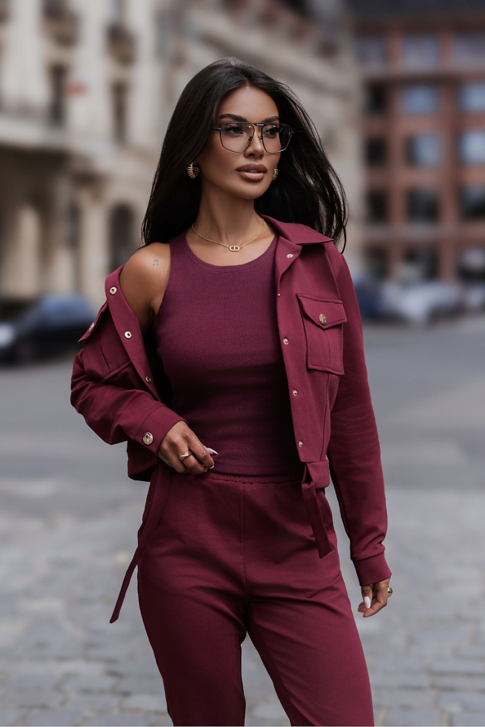 A woman walking outdoors on a city street wearing a matching burgundy outfit consisting of a cold-shoulder top, button-front jacket, and pants, along with glasses.