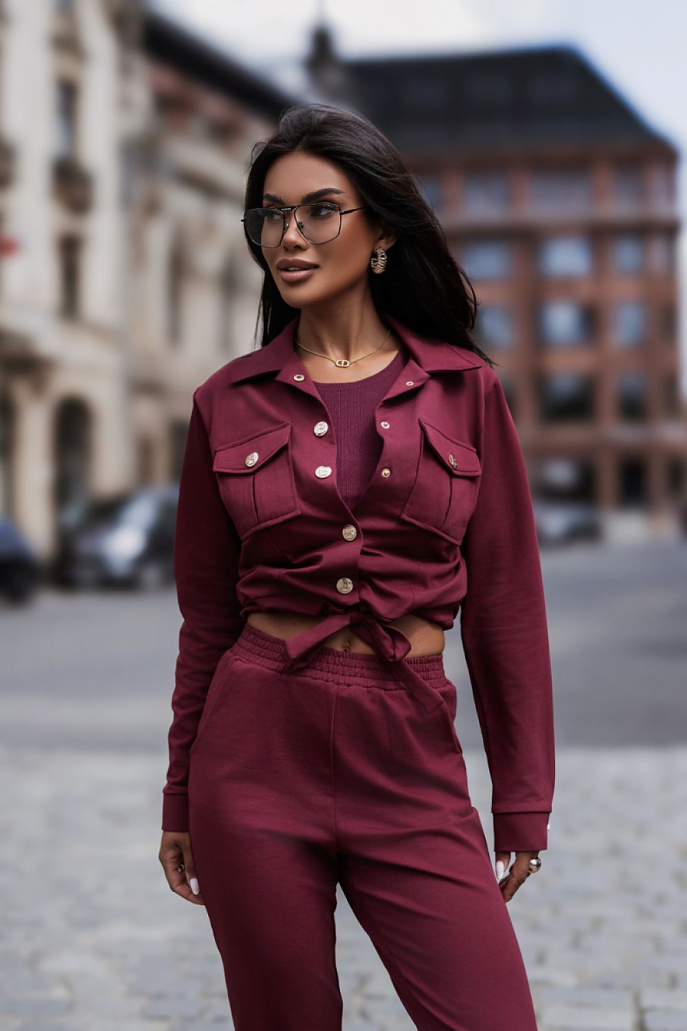 A woman wearing a matching burgundy two-piece set consisting of a cropped button-front jacket with chest pockets and high-waisted pants, standing outdoors on a cobblestone street with blurred buildings in the background.