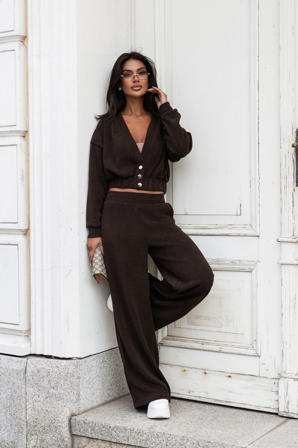 A woman models a dark brown two-piece lounge set consisting of a cropped long-sleeve cardigan with button details and matching wide-leg sweatpants, posing against a white paneled door.