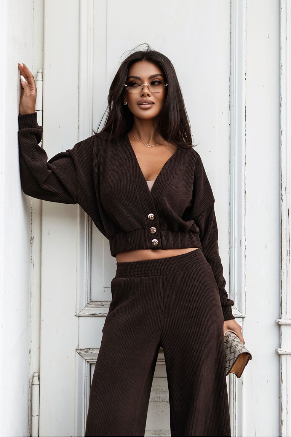 A woman wearing a dark brown ribbed knit two-piece set consisting of a cropped V-neck cardigan with buttons and matching wide-leg pants, posing against a white paneled door.