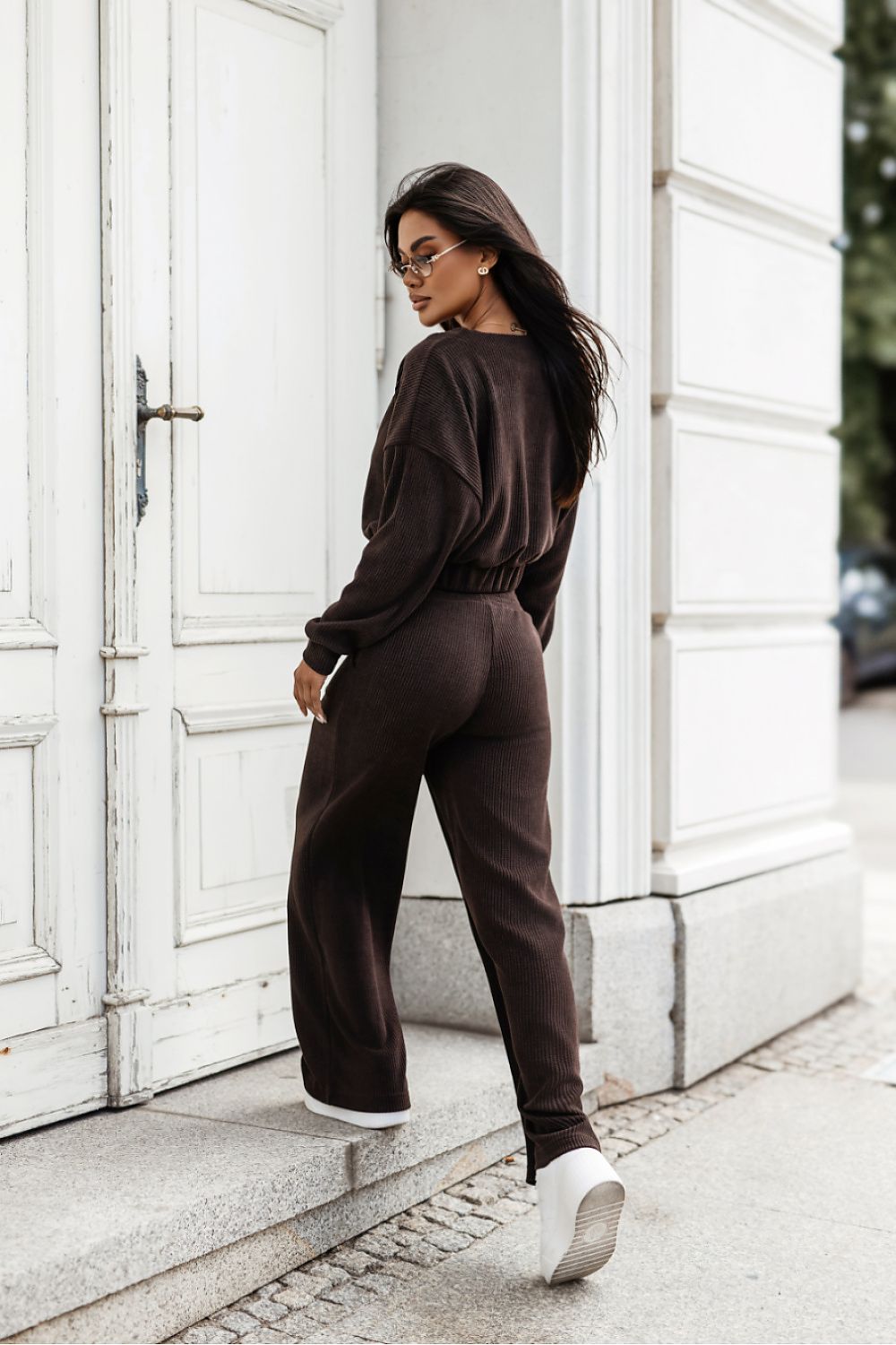 A woman wearing a matching dark brown lounge set consisting of a long-sleeve top and wide-leg pants, standing on stone steps in front of a white building.