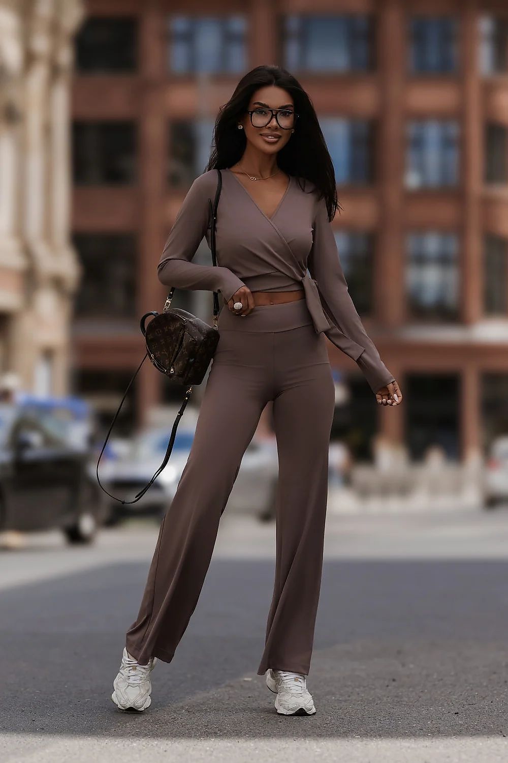 A woman wearing a matching taupe-brown two-piece lounge set consisting of a long-sleeve V-neck crop top and high-waisted wide-leg pants, styled with white sneakers and a dark patterned shoulder bag, standing on a city street.