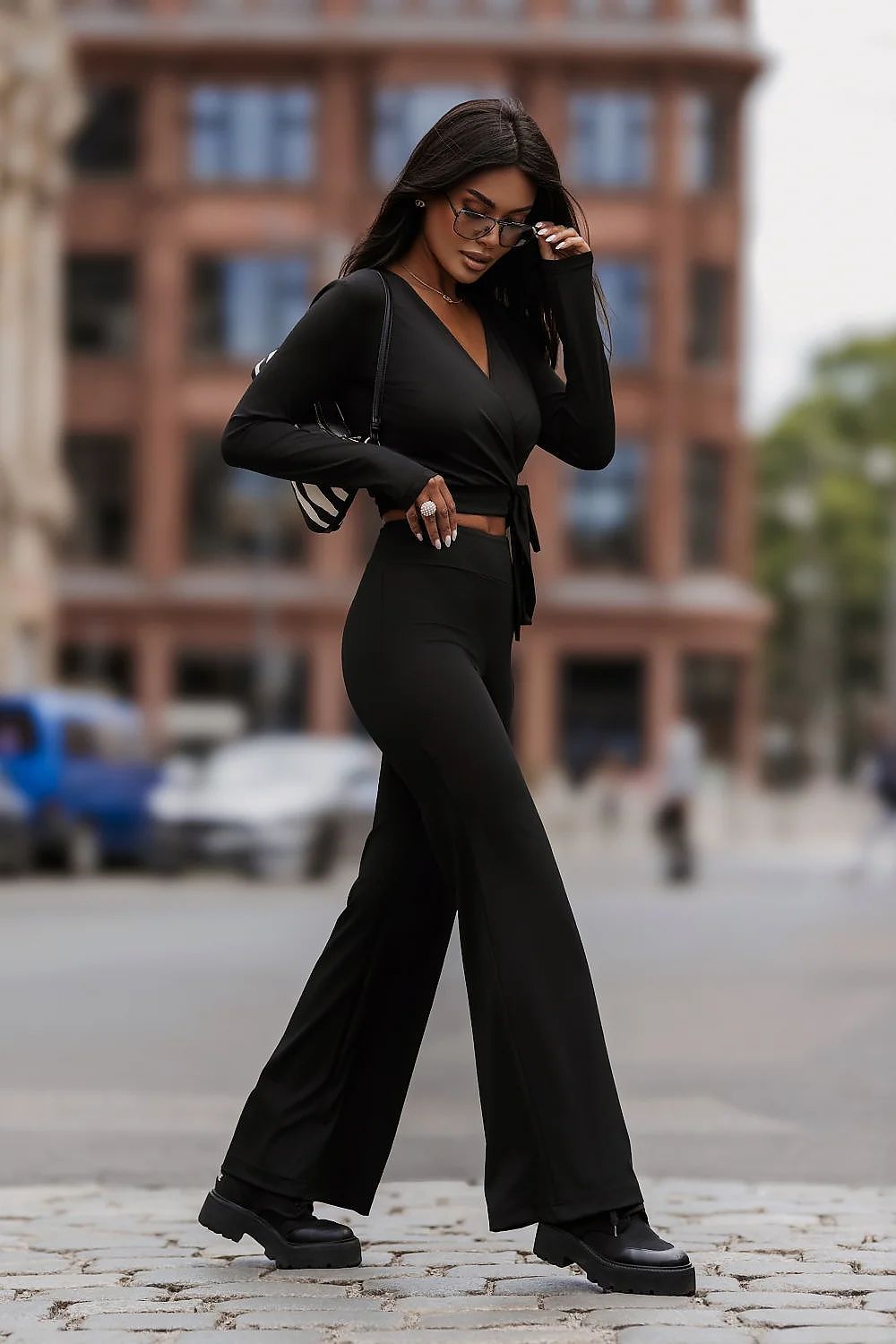 A woman wearing a black long-sleeve wrap-style jumpsuit with wide-leg pants and black platform shoes, walking on a cobblestone street with a blurred city background.