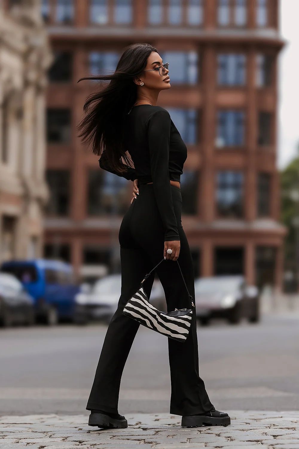 A woman walking outdoors in a black long-sleeve crop top and matching black pants, holding a black and white striped handbag, with a blurred city street and brick building in the background.