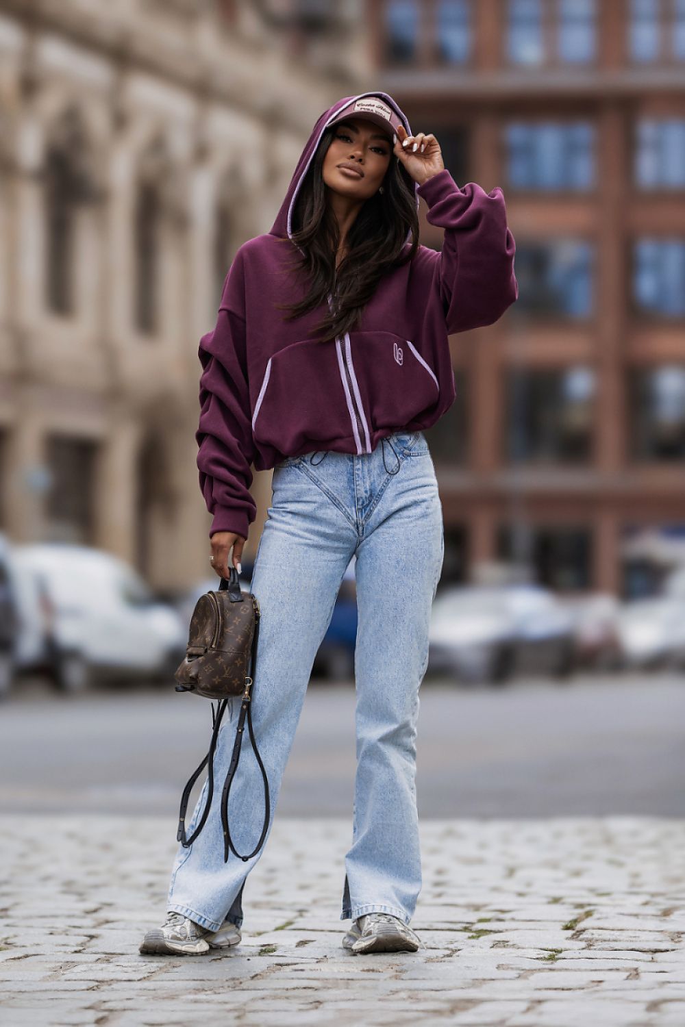 A woman stands on a cobblestone street wearing a maroon zip-up hoodie with white piping, light-wash wide-leg jeans, and carrying a small brown handbag.