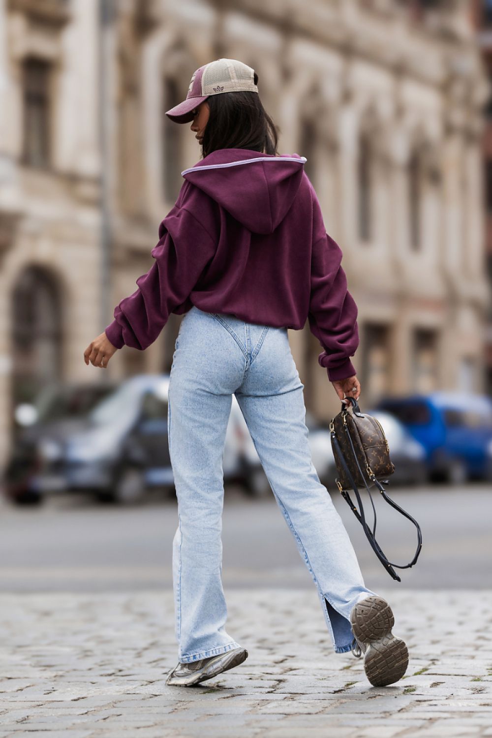 A woman walking on a cobblestone street, viewed from behind, wearing a deep purple cropped hoodie, light blue jeans, a beige baseball cap, and carrying a brown leather shoulder bag.