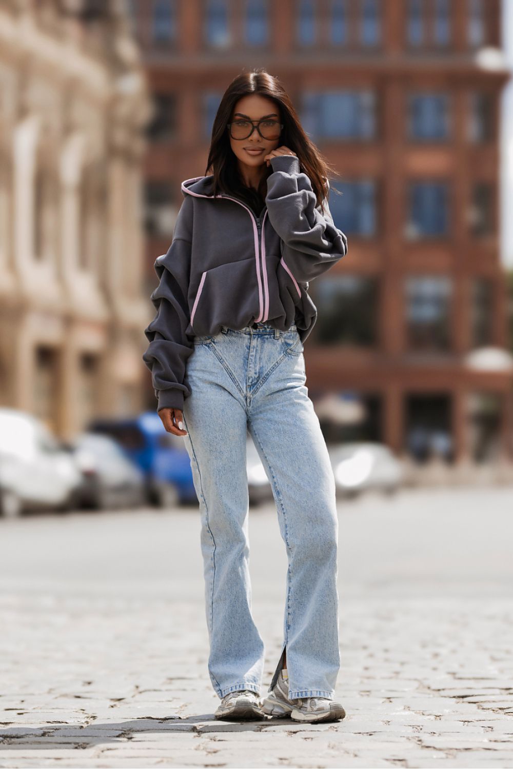 A woman stands on a cobblestone street wearing a dark gray zip-up hoodie with pink piping, light-wash blue jeans, and gray sneakers, with blurred buildings in the background.