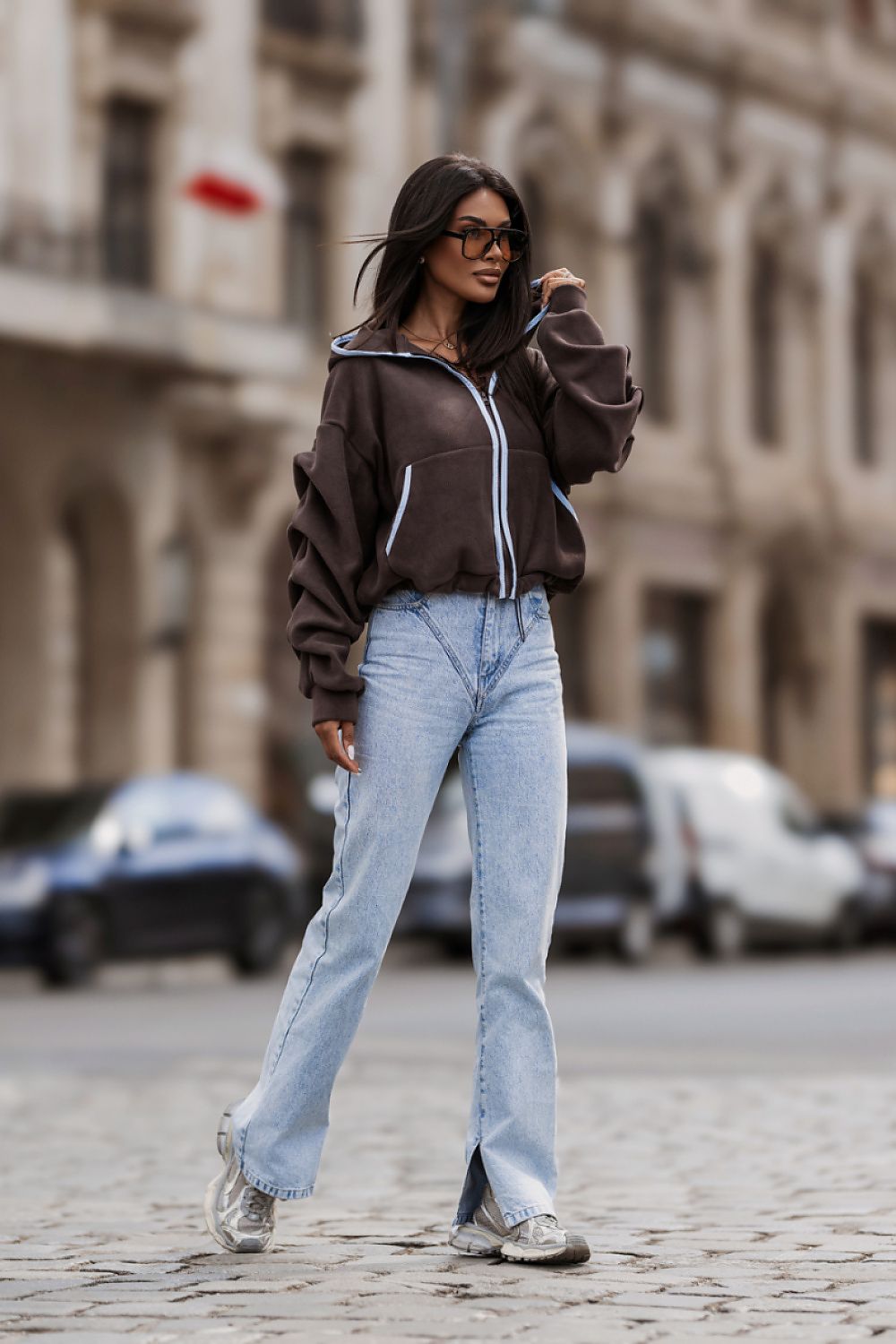 A woman stands on a cobblestone street wearing a dark brown cropped zip-up hoodie with light blue and white piping, light-wash blue jeans, and gray sneakers. The background shows a blurred city street scene with buildings and parked cars.