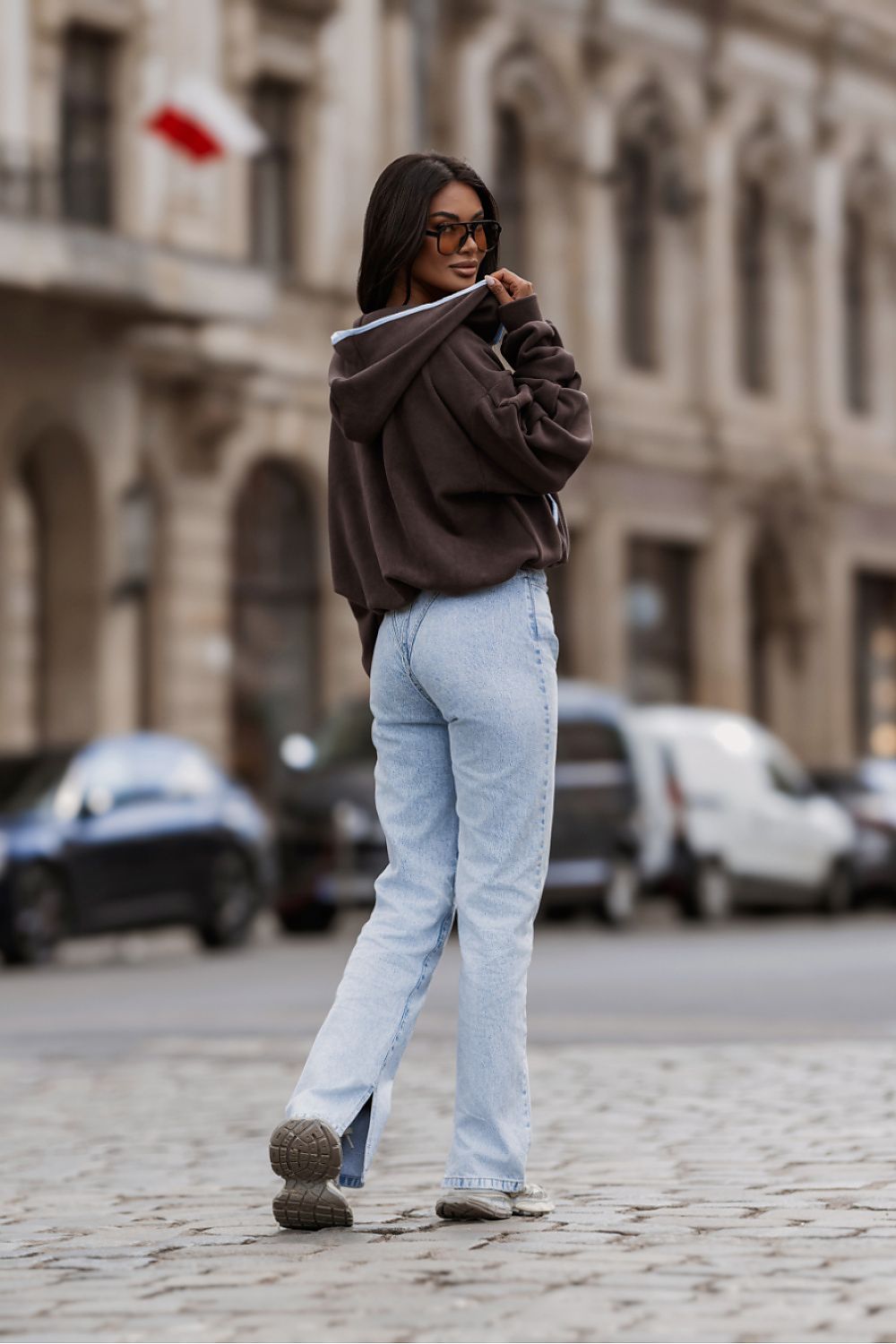 A woman wearing a dark brown cropped hoodie, light blue wide-leg jeans, and brown shoes walks on a cobblestone street in front of a blurred historic building.