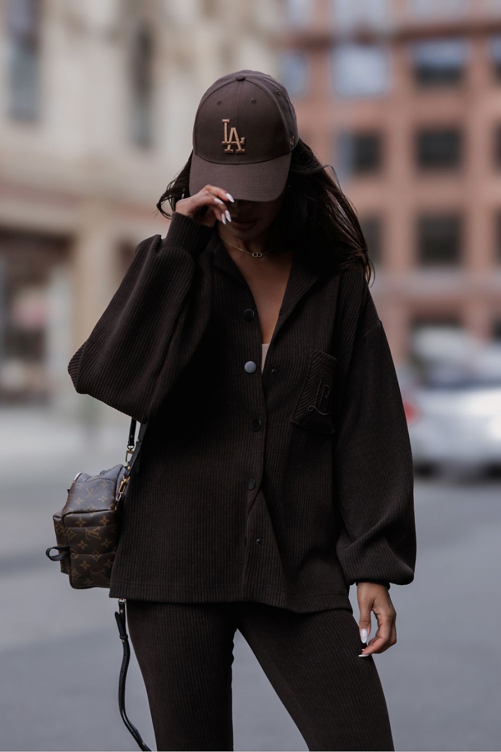 A person wearing a dark brown oversized button-up shirt, matching dark brown pants, a dark brown baseball cap with a gold 'LA' logo, and a brown patterned shoulder bag, standing on a city street.