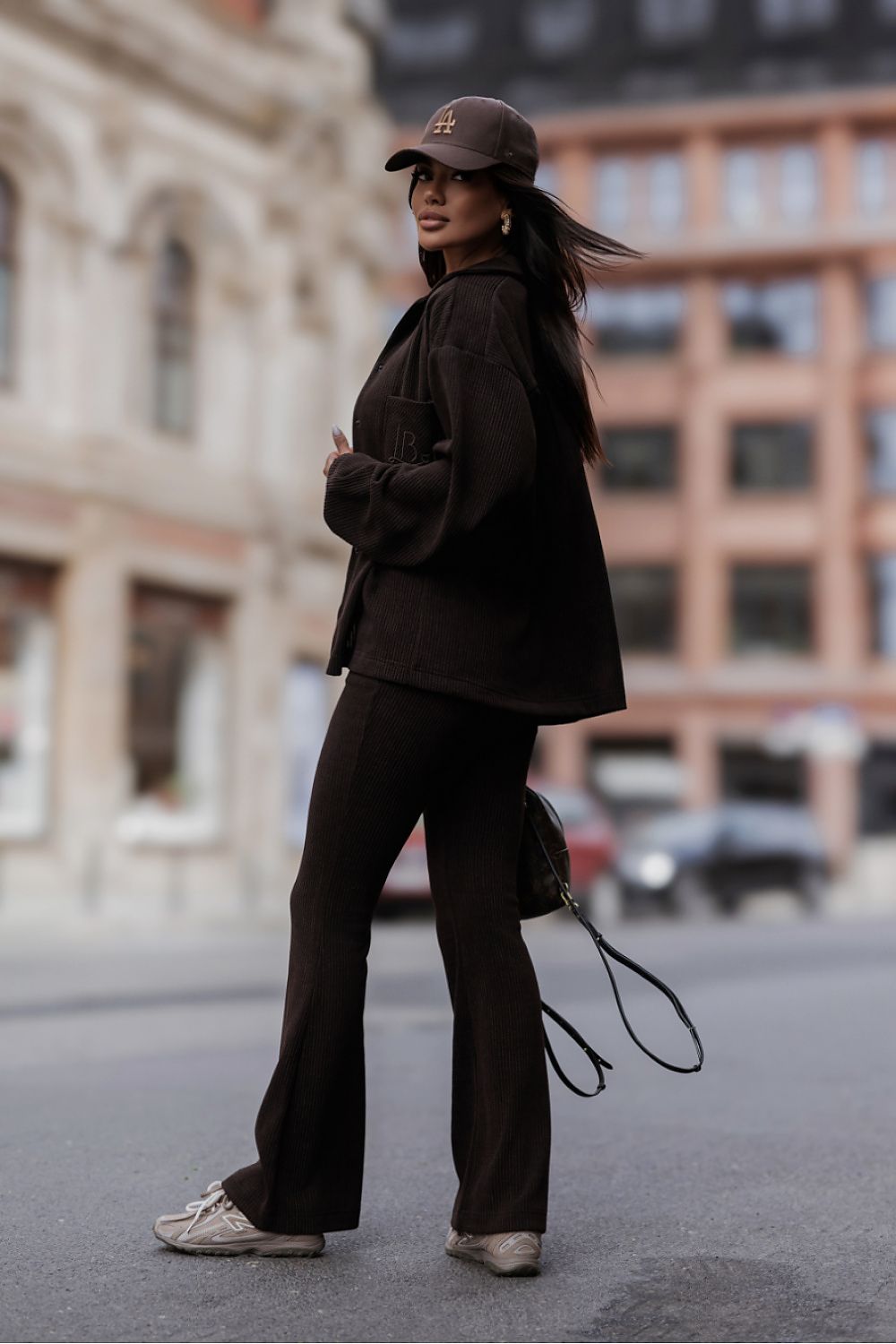 A woman stands on a city street wearing a dark brown oversized blazer, matching dark brown wide-leg pants, a dark brown baseball cap, and light-colored sneakers. A small dark shoulder bag is also visible.