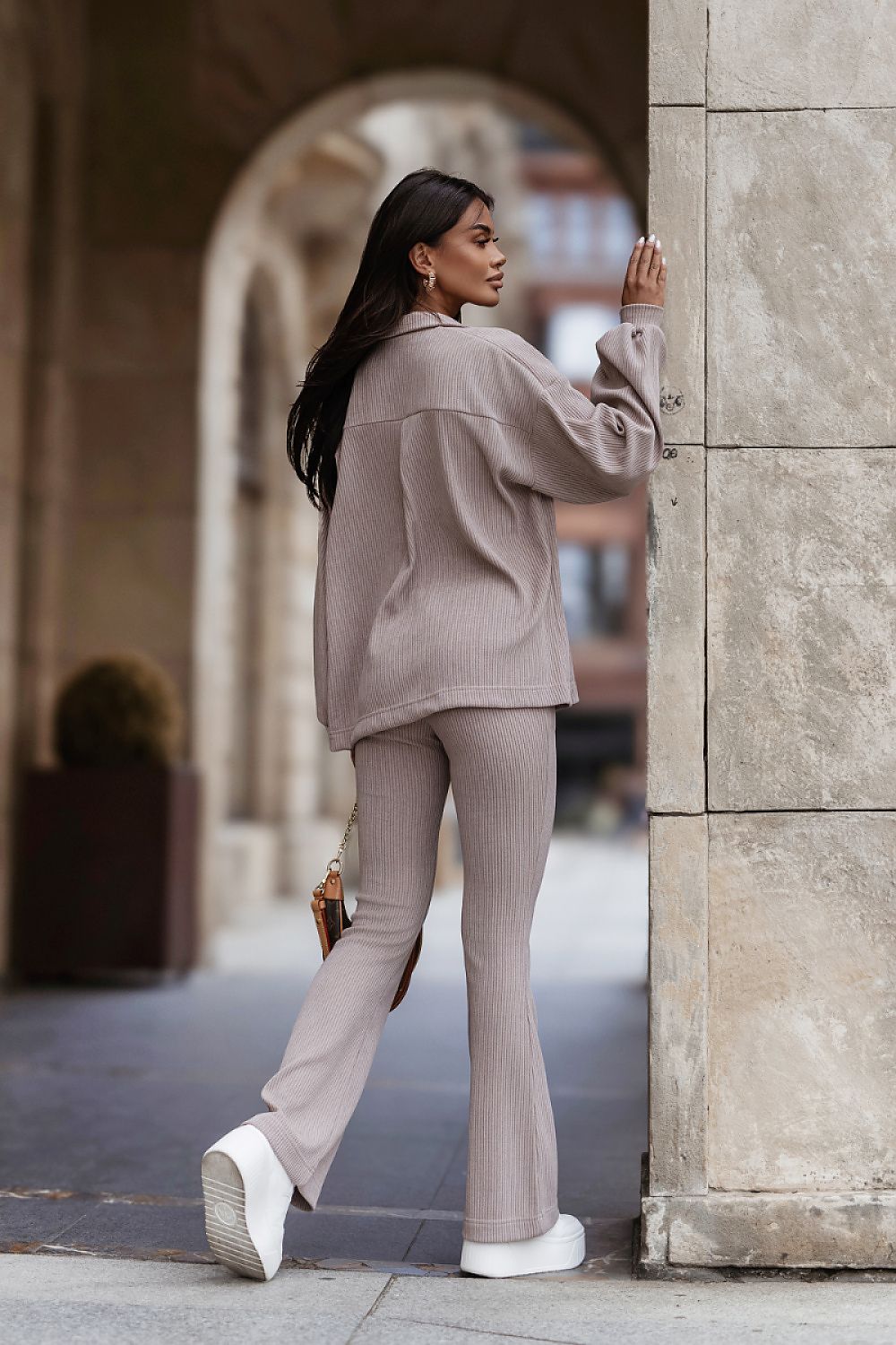 A woman posing in a matching taupe ribbed knit lounge set consisting of a cropped long-sleeve top and flared pants, standing in an outdoor stone walkway.