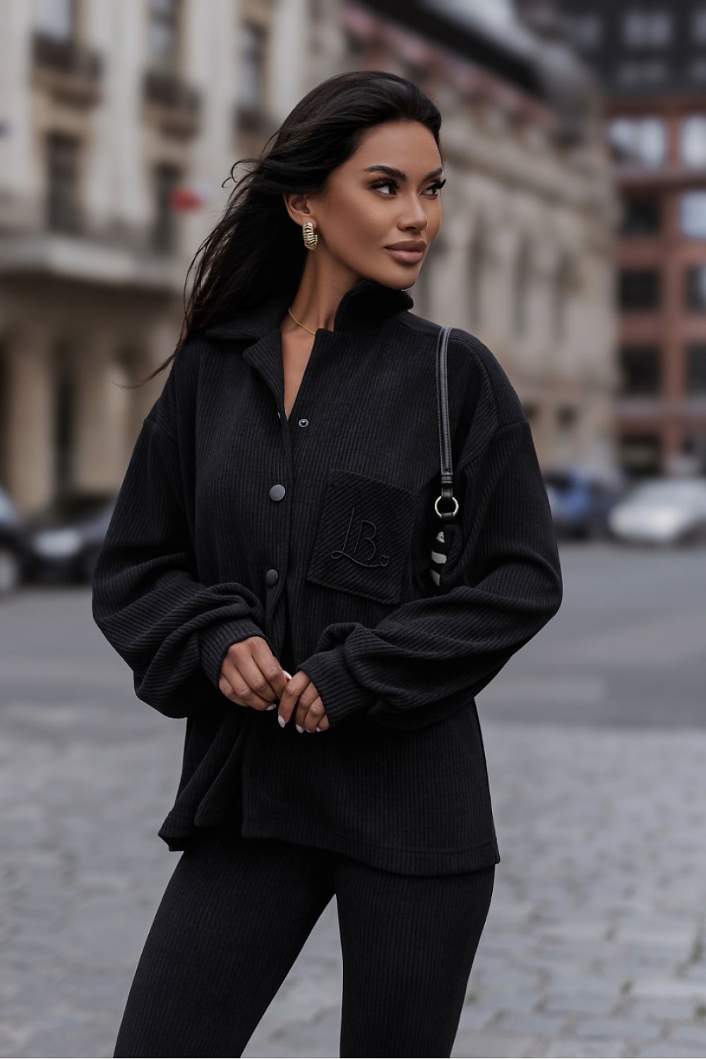 A woman wearing a black button-front overshirt with a chest pocket and black ribbed leggings, standing on a city street with buildings blurred in the background.