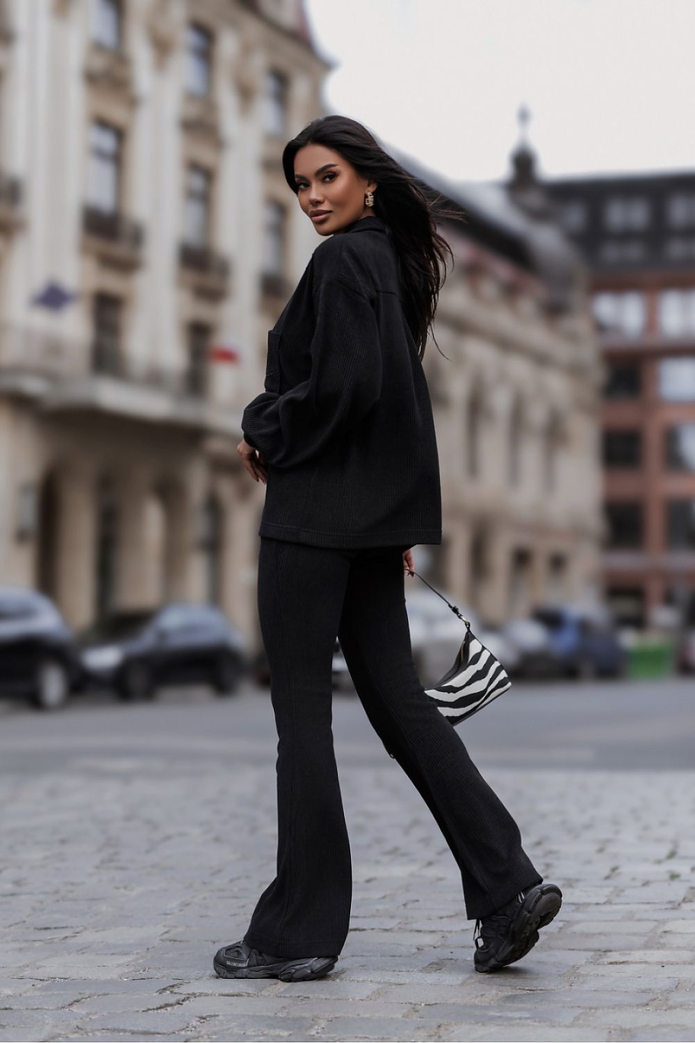 A woman walking on a cobblestone street wearing a black oversized long-sleeve top and matching black flared pants, carrying a black and white striped shoulder bag.