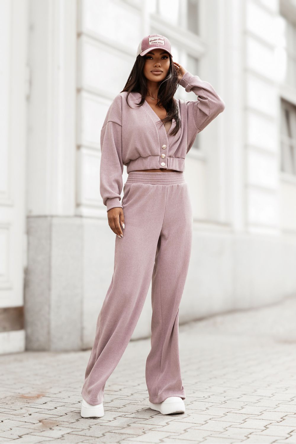 A woman stands outdoors on a paved walkway in front of a light-colored building, wearing a matching dusty mauve lounge set consisting of a cropped long-sleeve button-front top and wide-leg pants, along with a matching baseball cap and white sneakers.