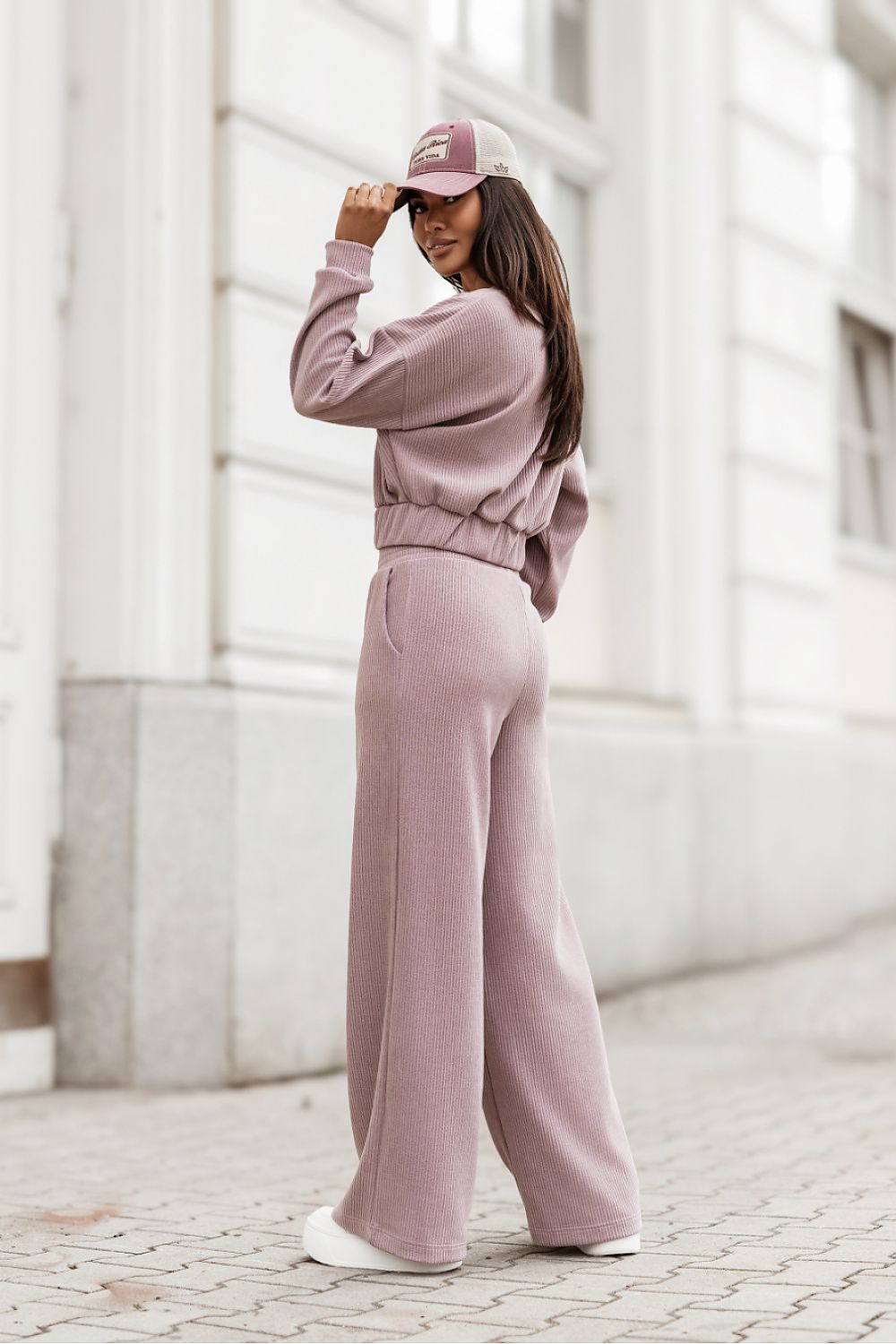 A woman wearing a dusty pink two-piece lounge set consisting of a cropped long-sleeve sweatshirt and matching wide-leg sweatpants, paired with a matching baseball cap and white sneakers, standing on a cobblestone street in front of a white building.