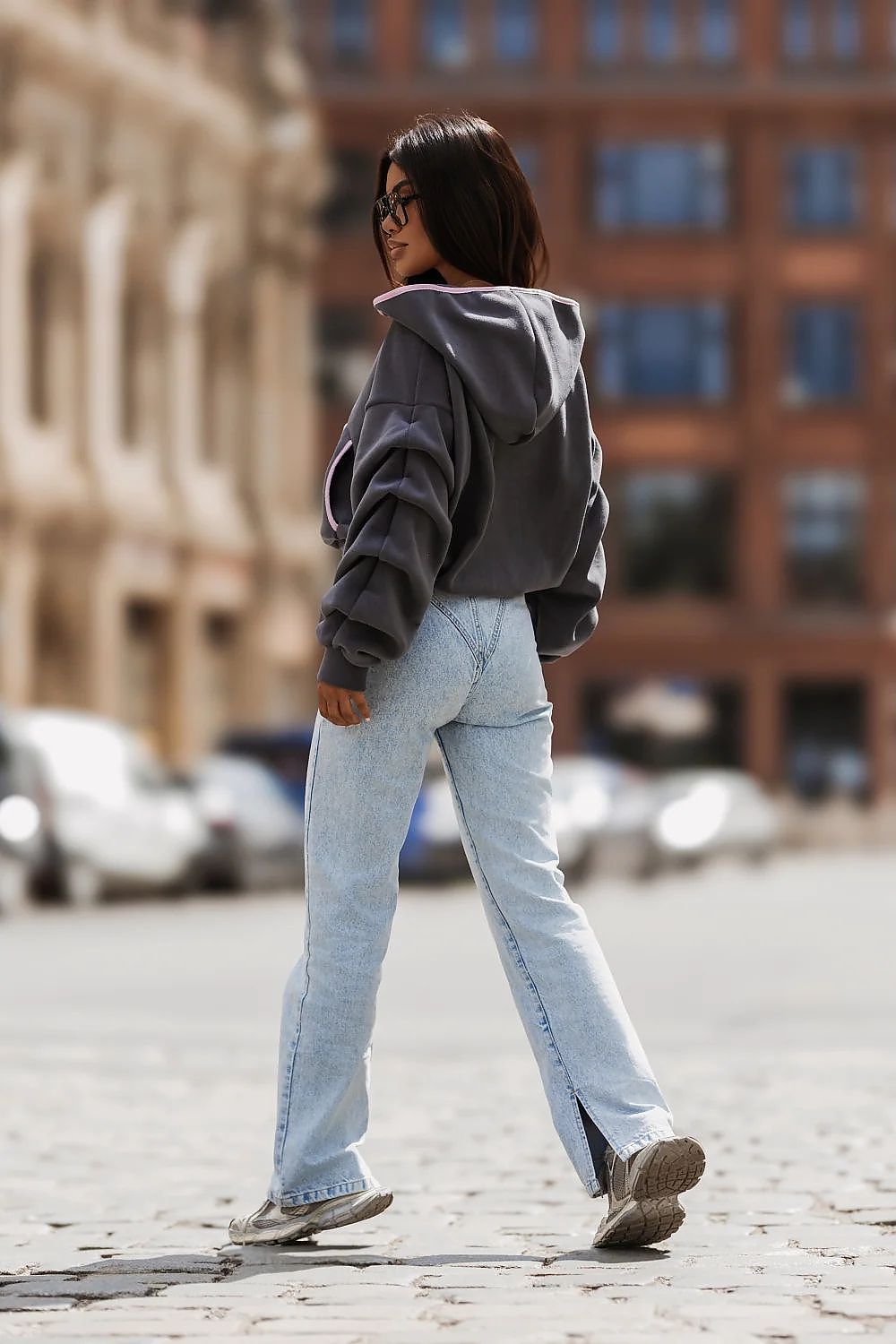 A woman walking outdoors on a cobblestone street wearing a dark gray cropped hoodie, light-wash wide-leg jeans, and brown sneakers, with blurred buildings in the background.
