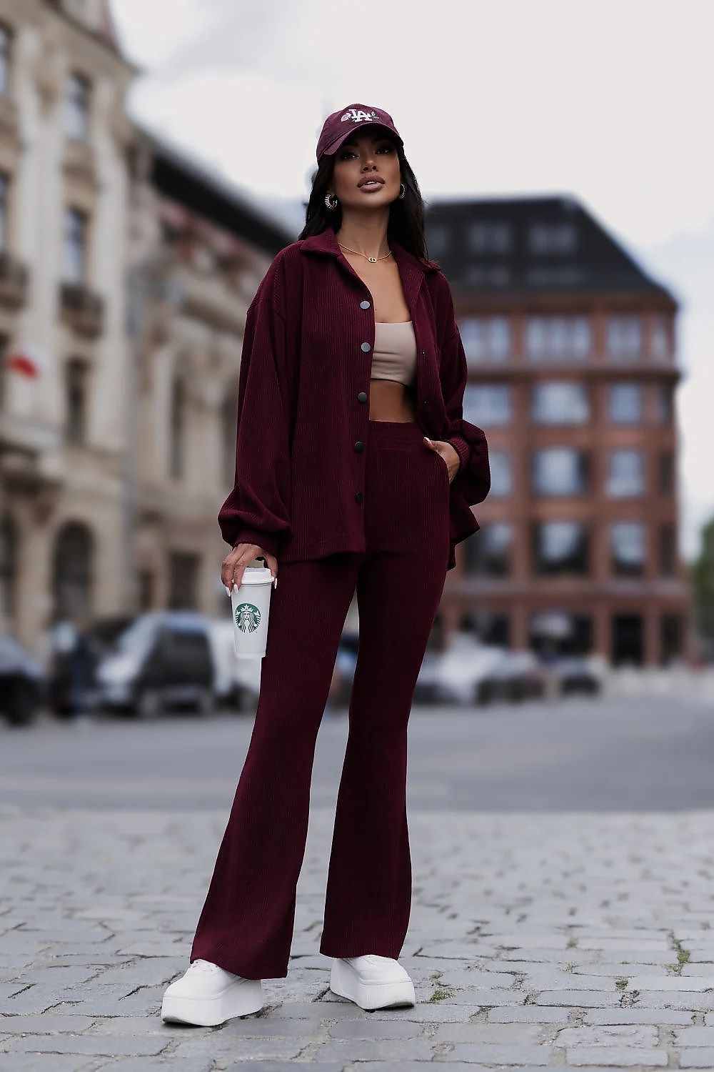 A woman stands on a cobblestone street wearing a matching burgundy two-piece outfit with a button-front overshirt and wide-leg pants, a burgundy baseball cap, a beige crop top, white sneakers, and holding a Starbucks cup.