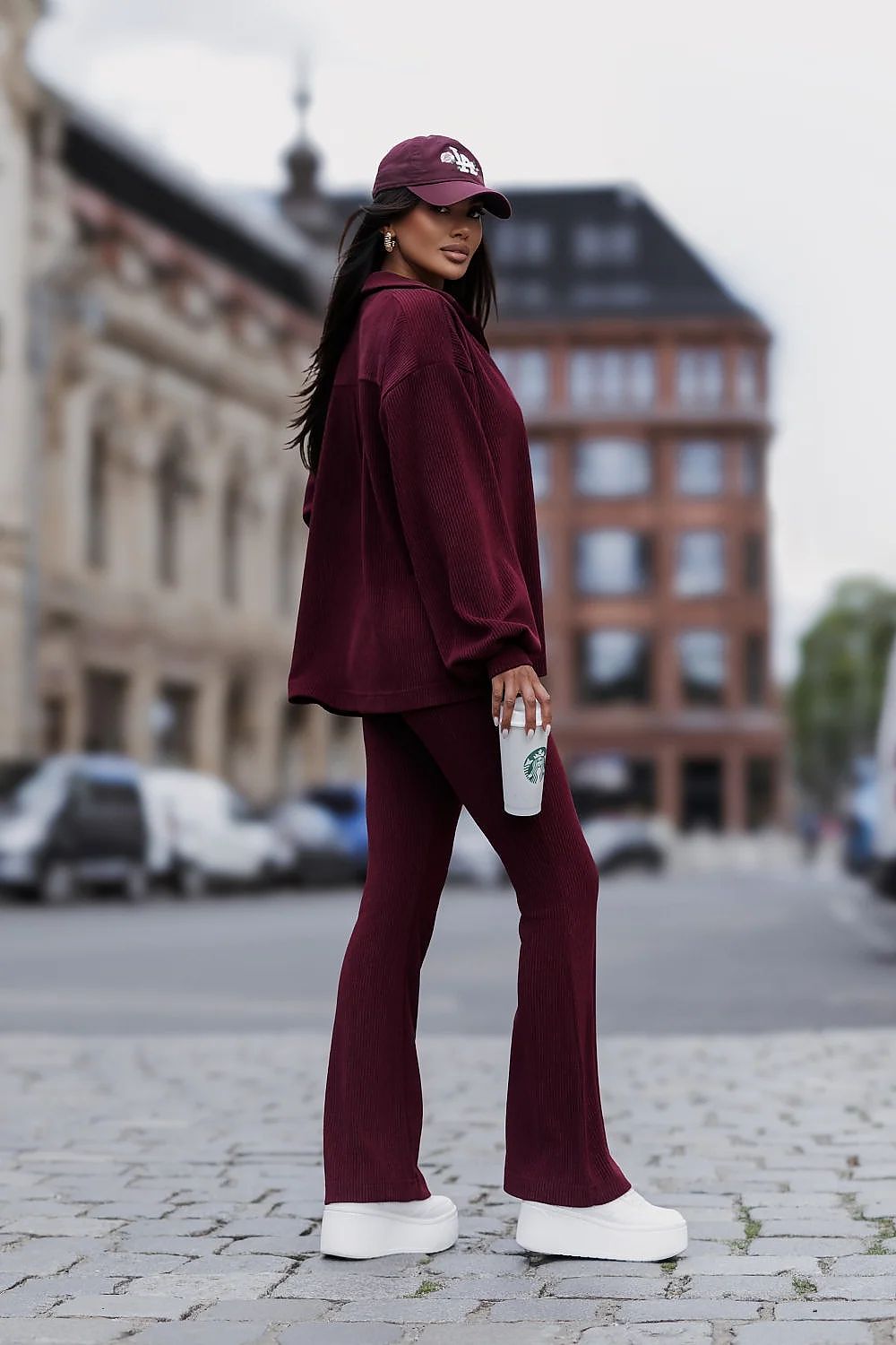 A woman stands on a cobblestone street wearing a matching burgundy lounge set consisting of a long-sleeve sweatshirt and wide-leg pants, a burgundy baseball cap, and white platform sneakers. She is holding a white Starbucks cup.