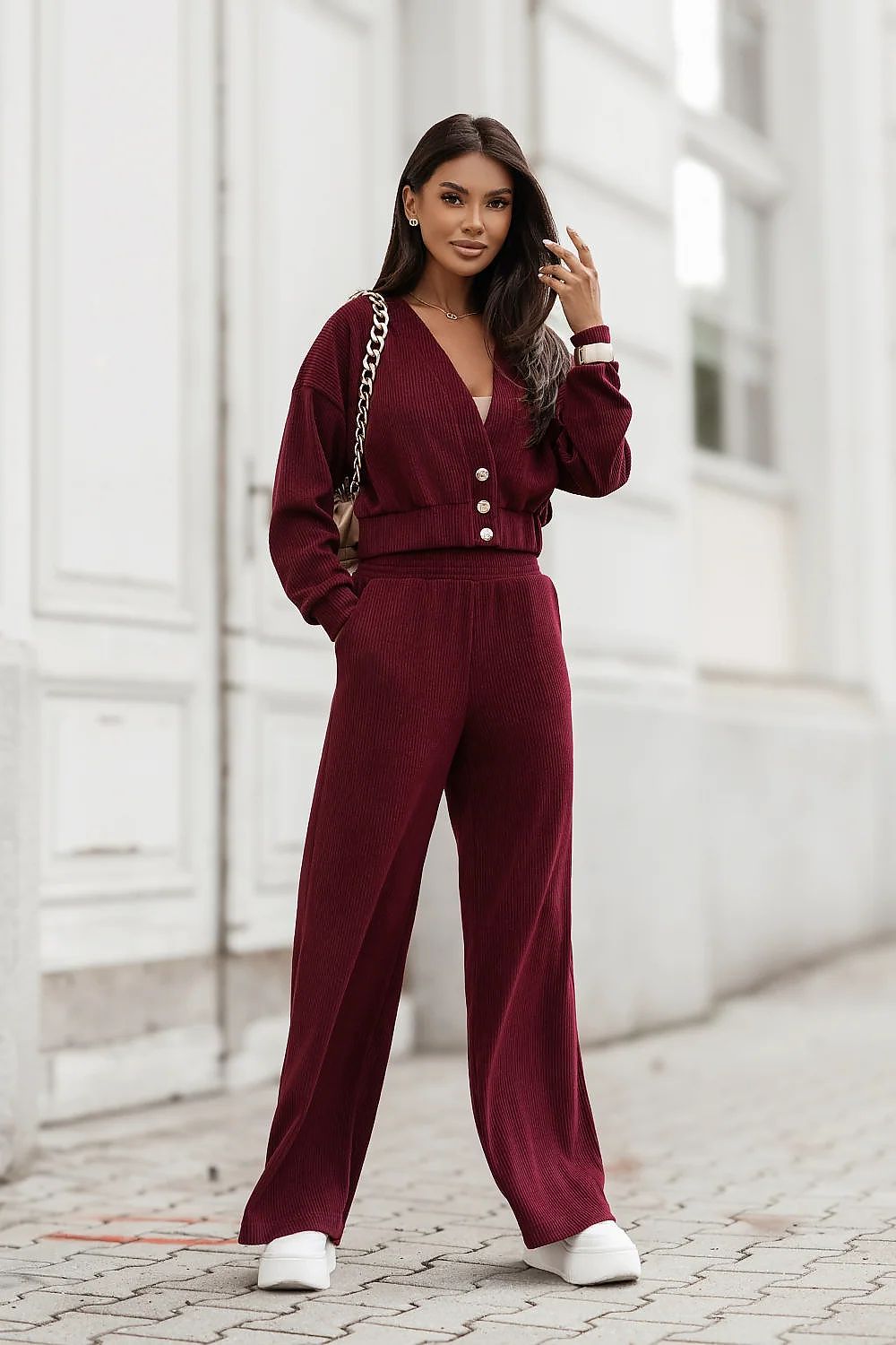 A woman stands outdoors on a paved street in front of a white building, wearing a matching burgundy long-sleeve top and wide-leg trousers set with white buttons at the waist. She is also wearing white sneakers and carrying a shoulder bag.