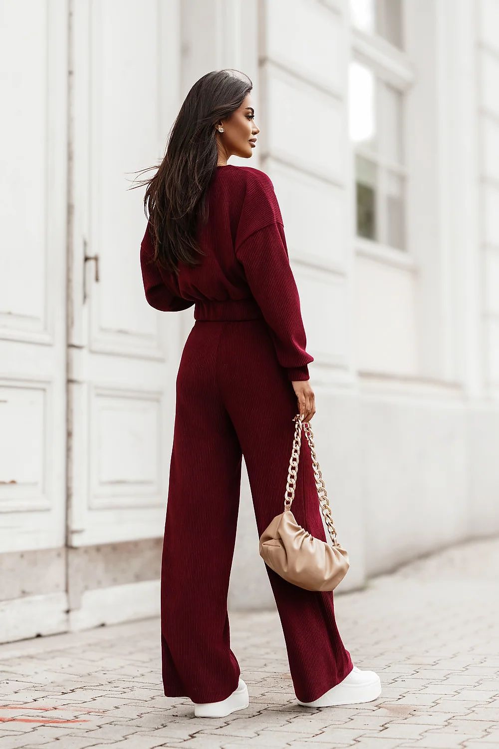 A woman stands on a cobblestone street in front of a white building, wearing a deep burgundy wide-leg jumpsuit and holding a beige handbag with a gold chain strap.