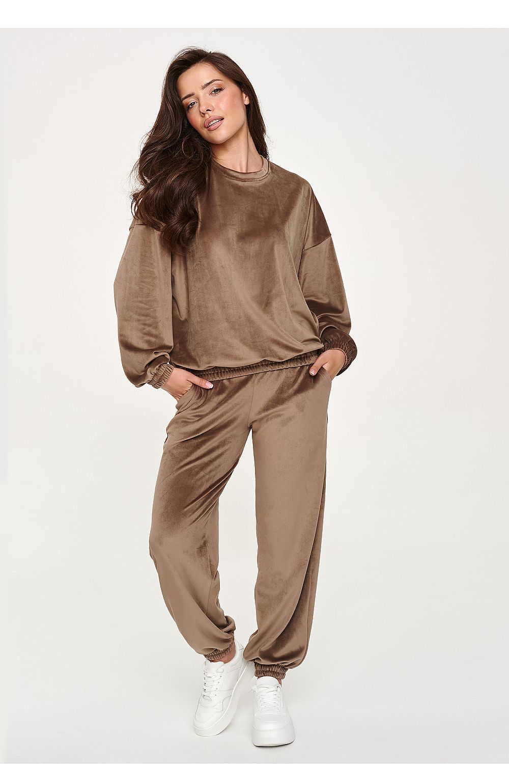 A woman modeling a matching brown velvet lounge set consisting of a long-sleeve sweatshirt and jogger pants, paired with white sneakers, against a plain light gray background.