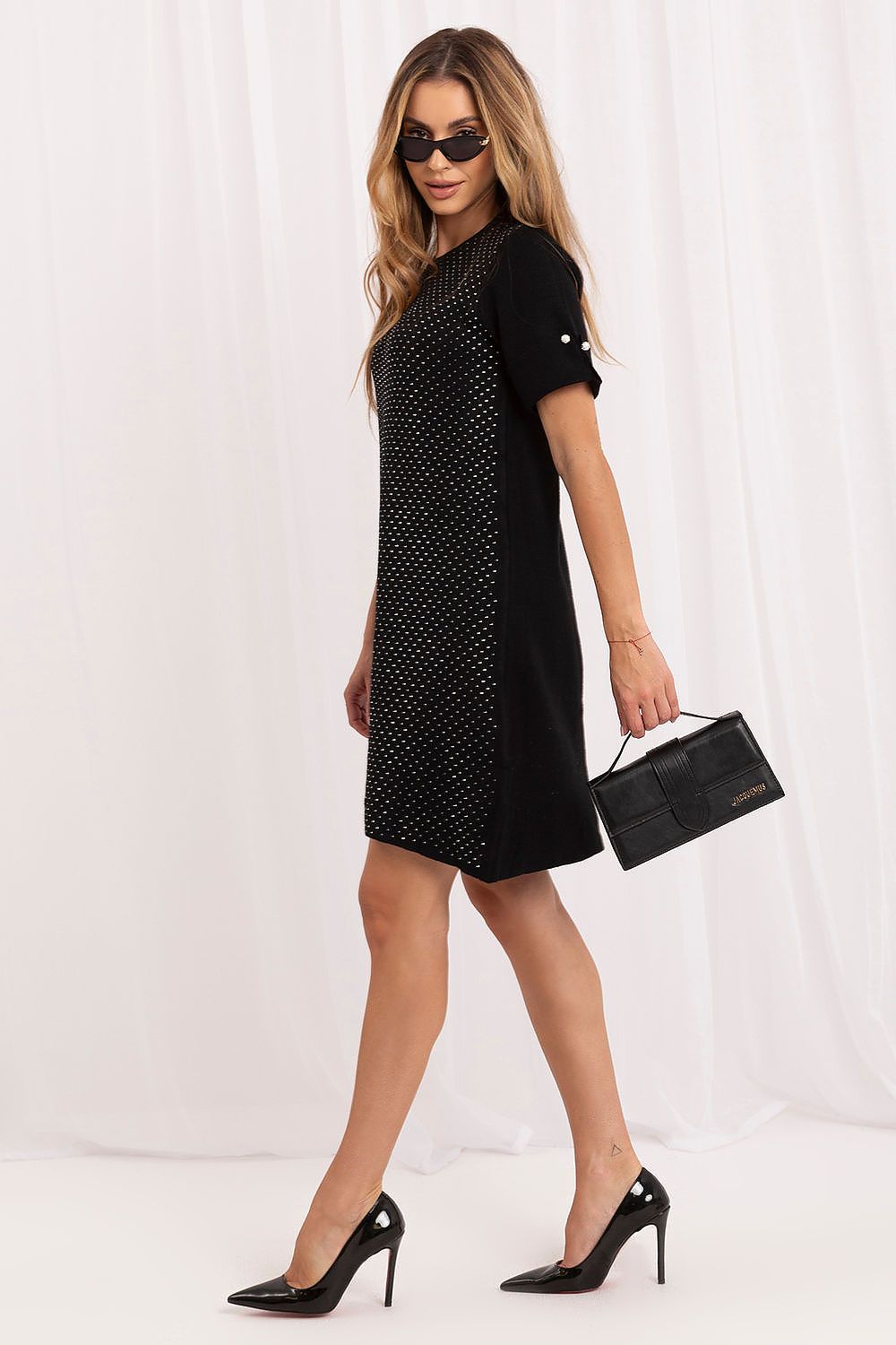 A woman models a short-sleeved black dress with a textured dotted pattern on the front panel, styled with black pointed-toe heels, sunglasses, and a black handbag against a white draped studio backdrop.