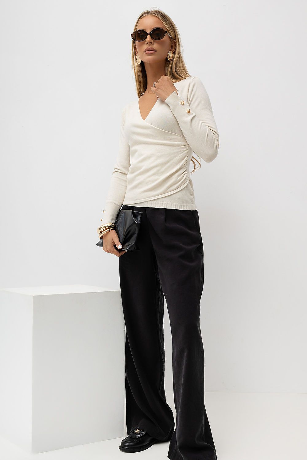 A woman modeling a cream wrap-style long-sleeve top and black wide-leg trousers, standing in a bright studio with a white block pedestal.