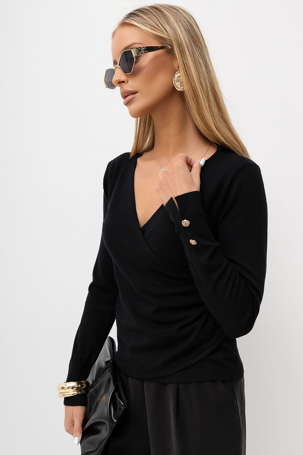 A woman wearing a black long-sleeve wrap-style top with gold buttons on the sleeve, styled with black bottoms, sunglasses, and a black clutch bag against a light gray background.