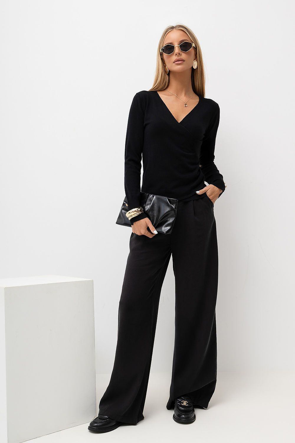 A woman modeling a black long-sleeve wrap top and matching wide-leg trousers, styled with black sunglasses, a black clutch, and black loafers with gold hardware, standing against a white studio background.