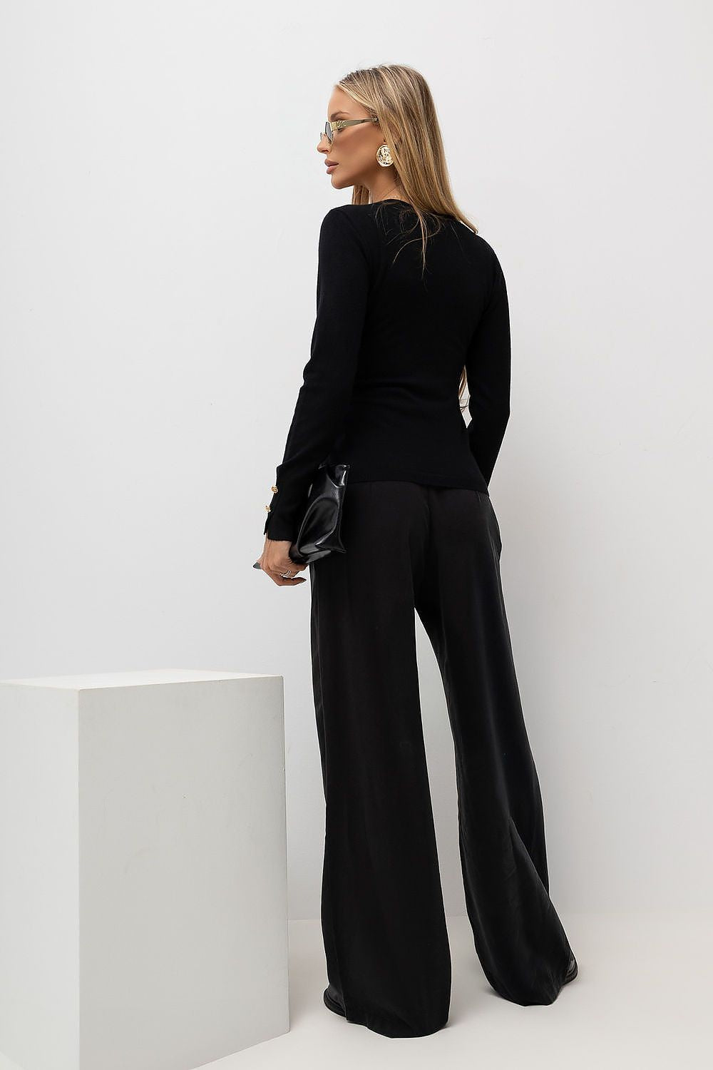 A woman stands in profile wearing a black long-sleeve top and wide-leg black pants, holding a small black clutch, against a clean white studio background with a white pedestal.