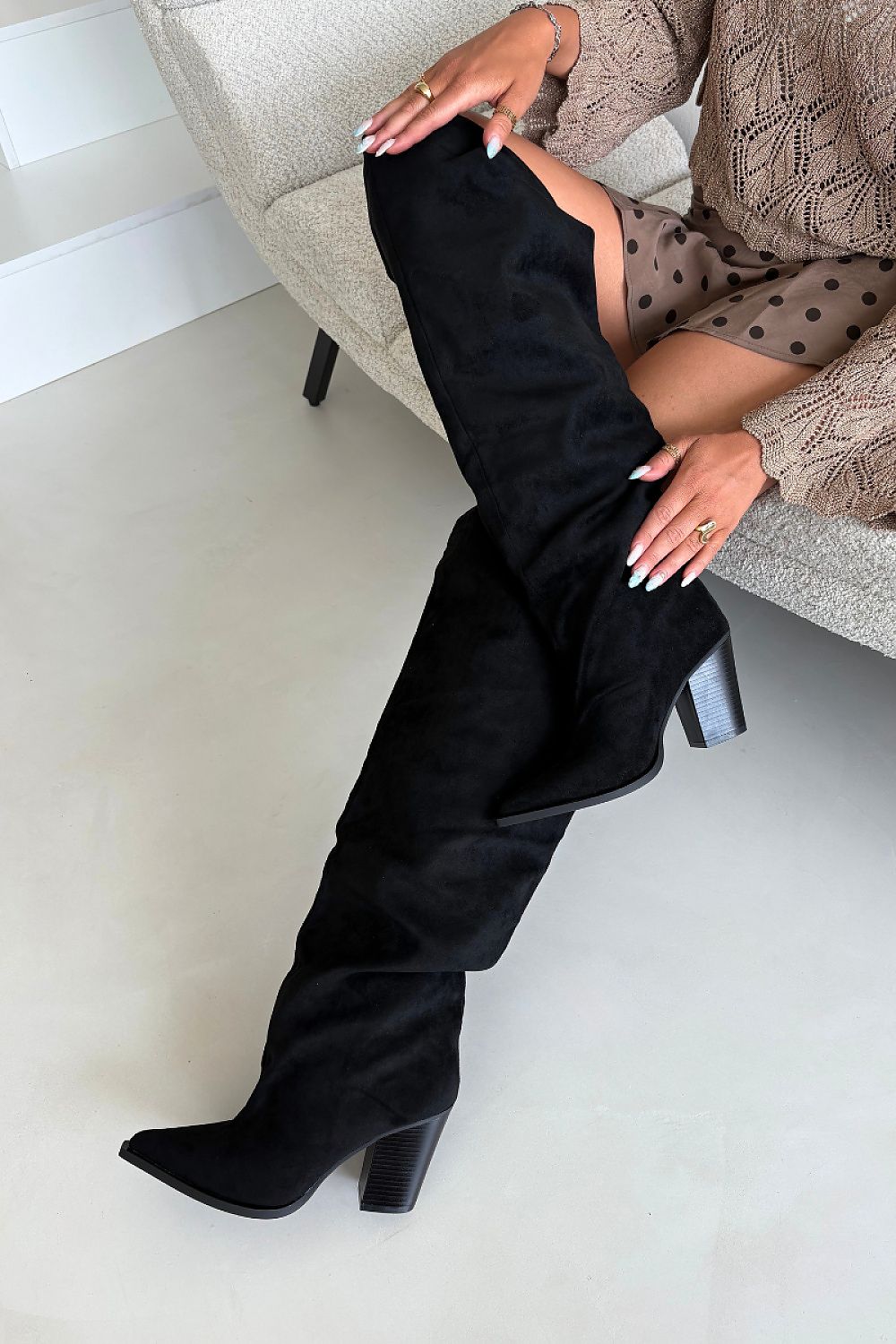 A person seated on a light beige sofa wearing a long black over-the-knee boot with a block heel. The photo is cropped to show the lower body, hands, and part of the outfit in an indoor room setting.