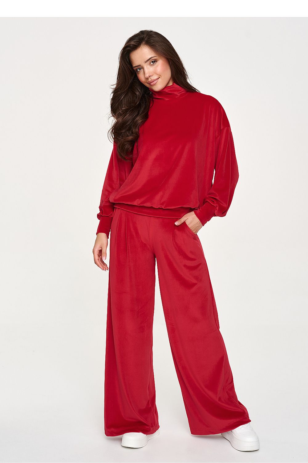 A woman modeling a matching red two-piece lounge set consisting of a long-sleeve mock-neck top and wide-leg pants, styled with white sneakers against a plain light gray studio background.