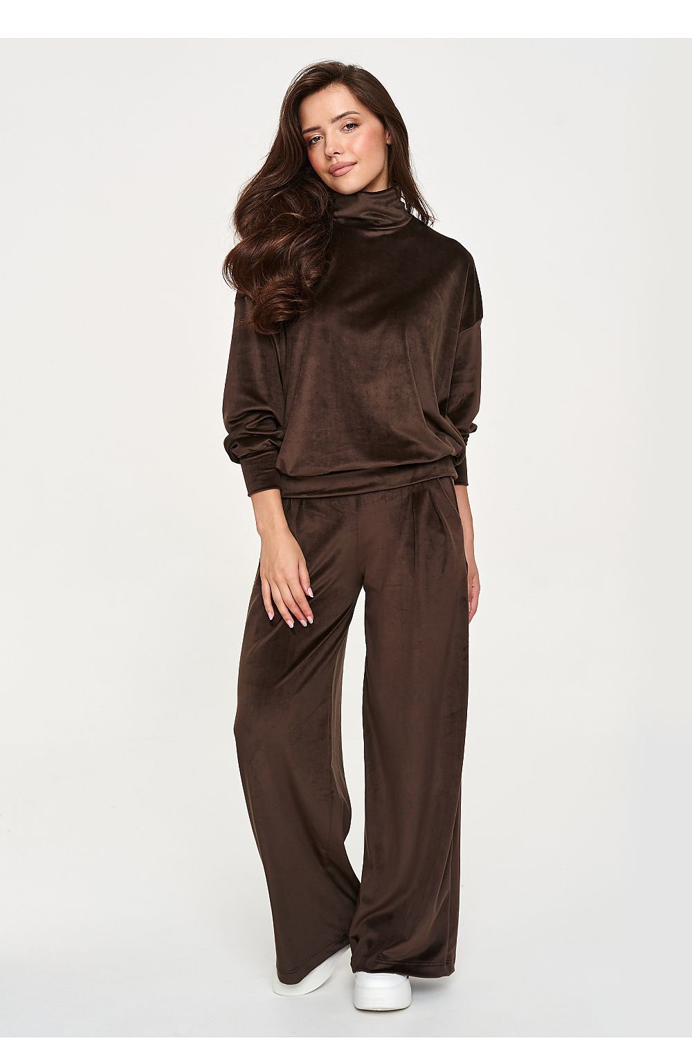 A woman modeling a matching dark brown velour lounge set consisting of a long-sleeve mock neck top and wide-leg pants, paired with white sneakers, against a plain light gray background.